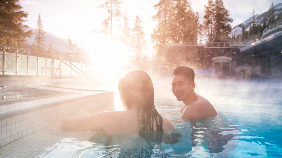 Smiling couple soaks in Banff Upper Hot Springs as steam rises from the geothermal waters