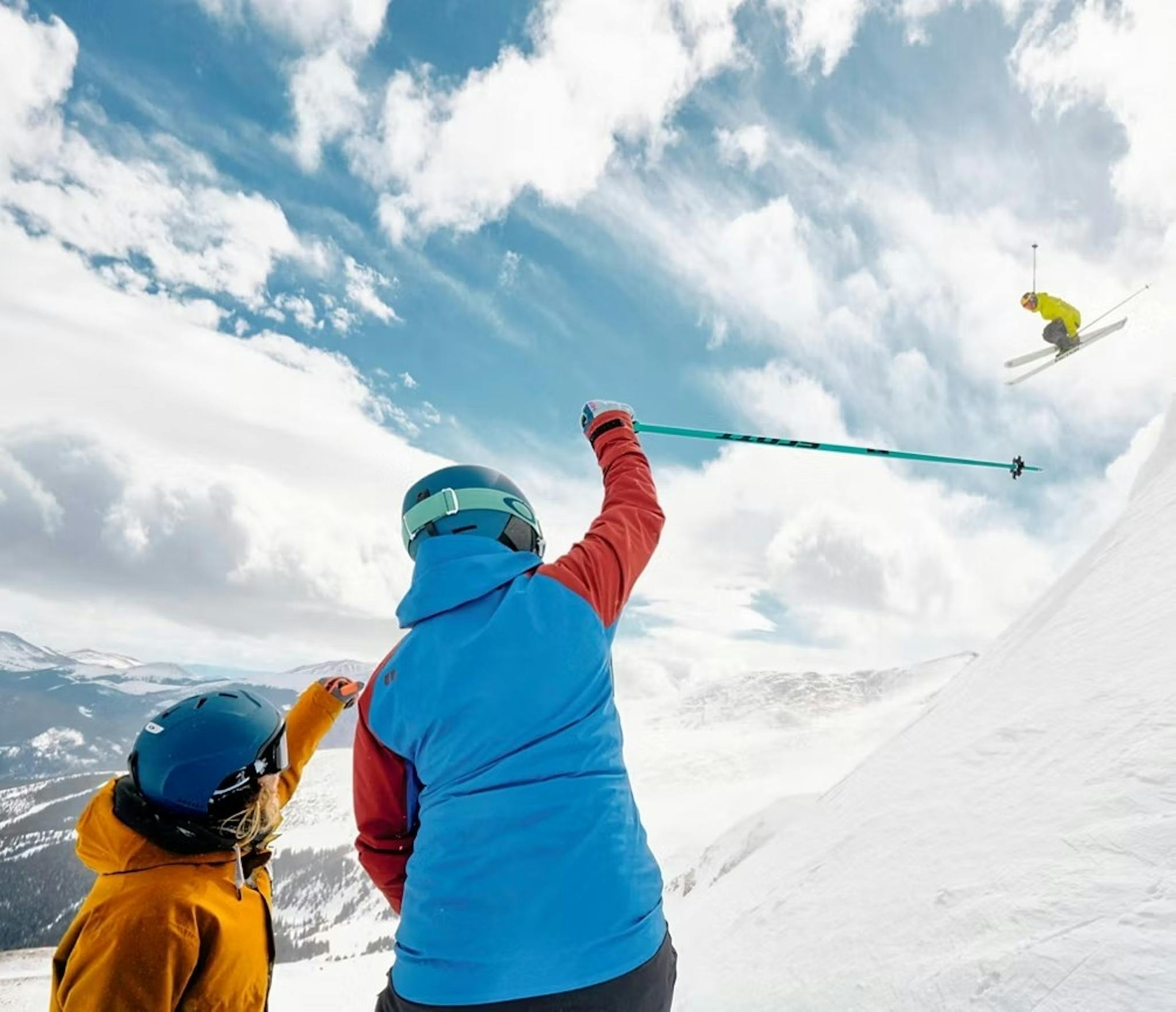 Two skiers watching and cheering on another skier in a yellow ski jacket perform a jump off a snowy mountain ridge at Breckenridge in Colorado, USA, in May.