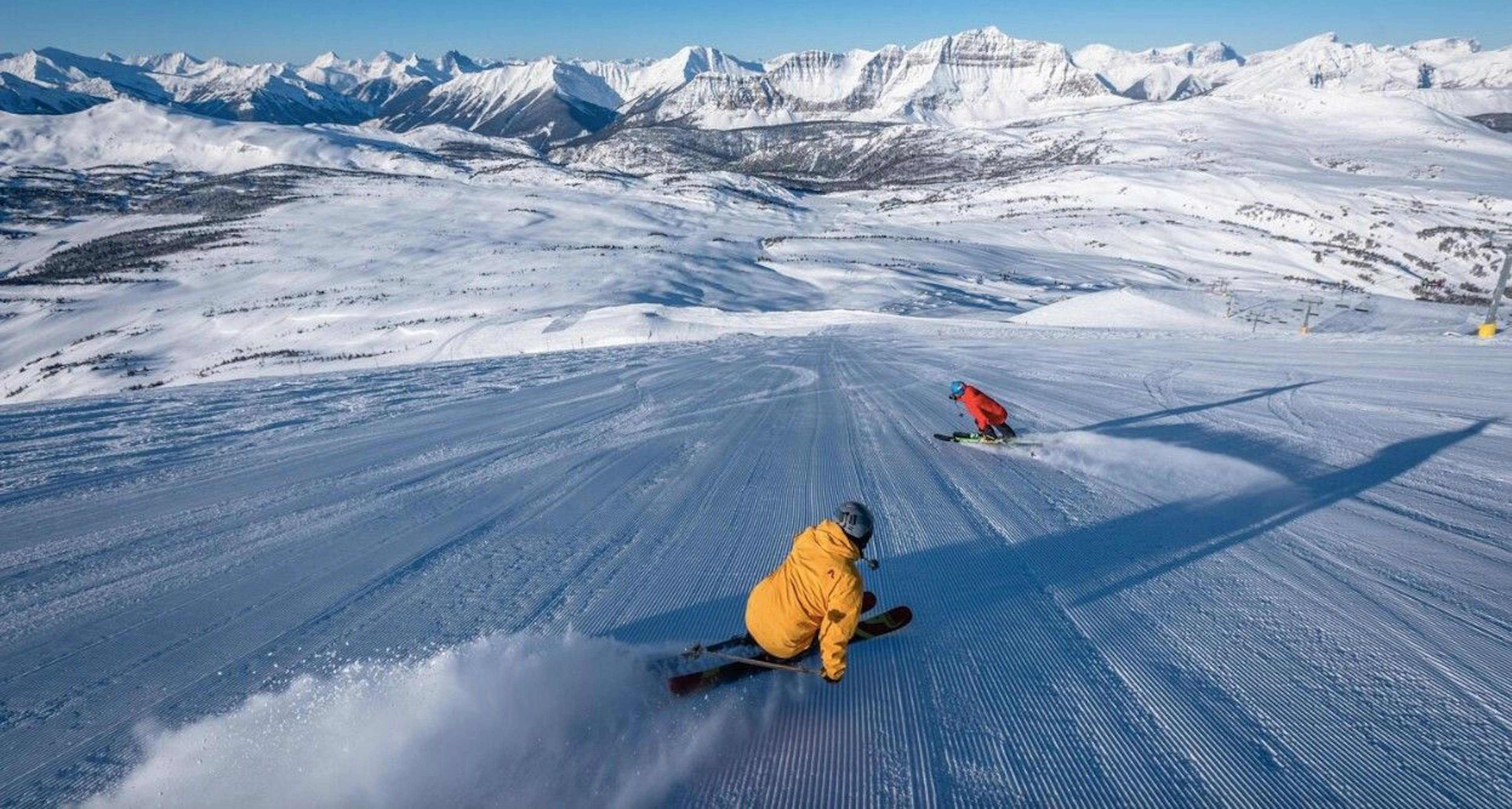 Two skiers going downhill on a groomed slope overlooking snowy mountains on a sunny day at Banff Lake Louise Ski Resort