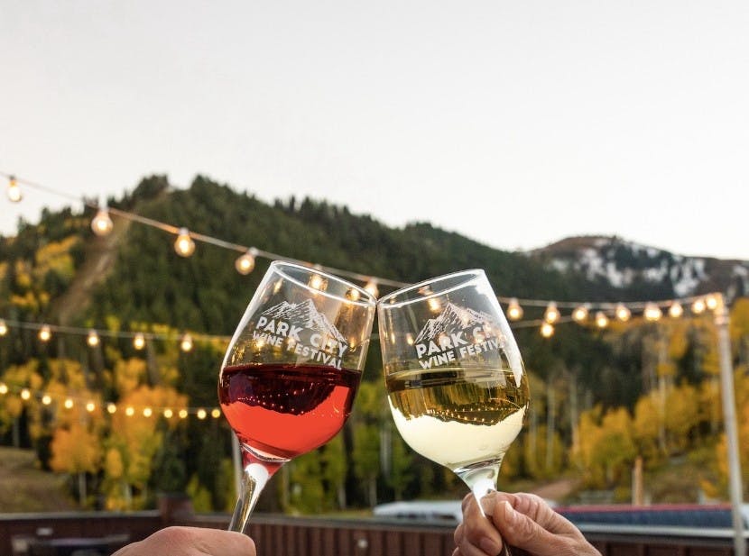 Two hands holding clinking wine glasses that read Park City Wine Festival with lights and mountains in the backdrop