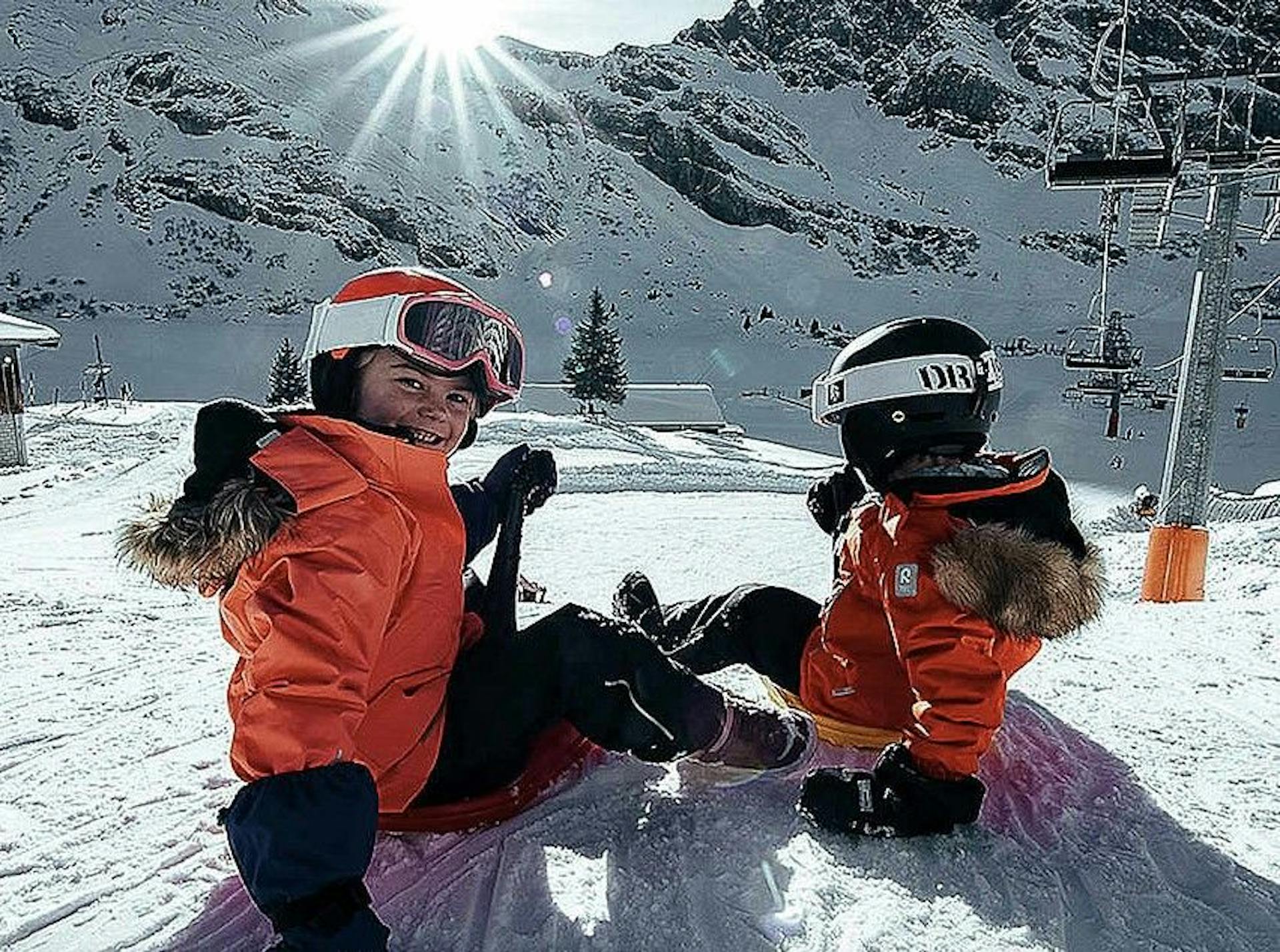 2 smiling kids in ski gear sit in the snow on a sunny day