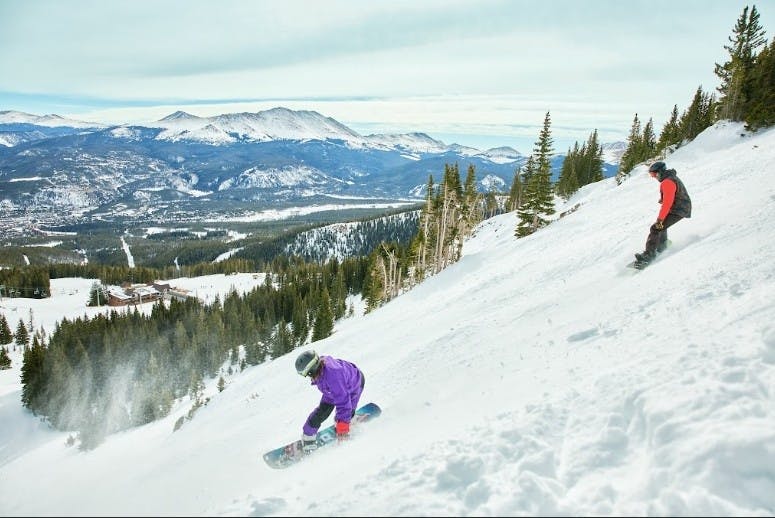 two snowboarders going down a run at Breckenridge with a snowy mountain backdrop
