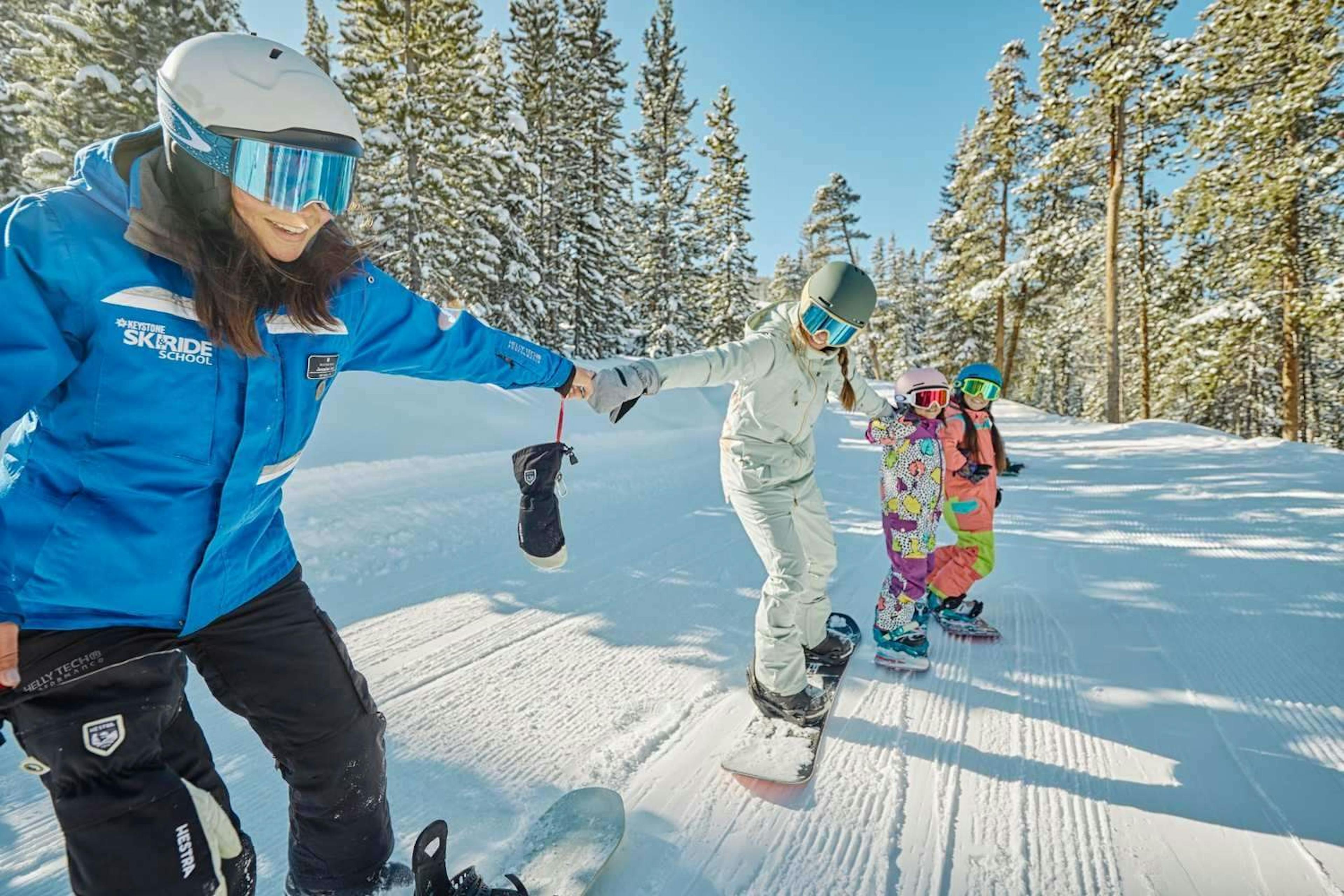 a snowboard instructor wearing a blue coat takes small kids on a ski track at keystone mountain resort. 