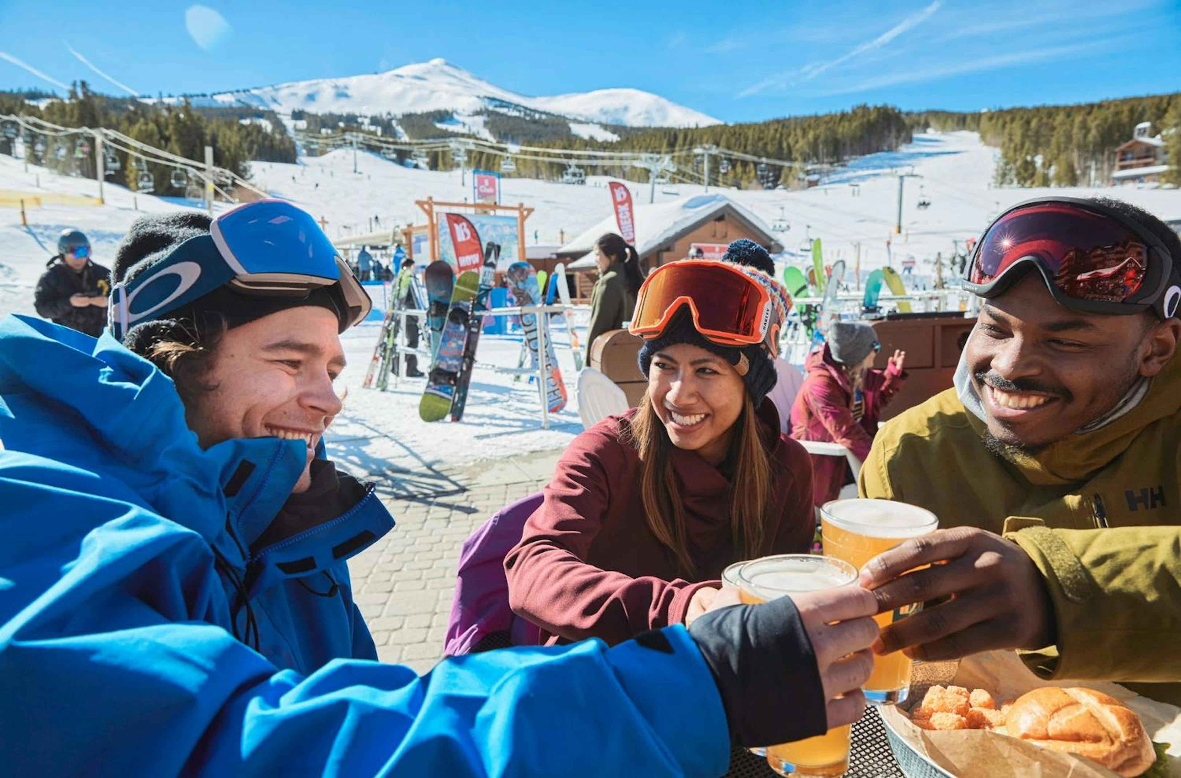 Three skiers clanking beer glasses at the base of Breckenridge ski resort.