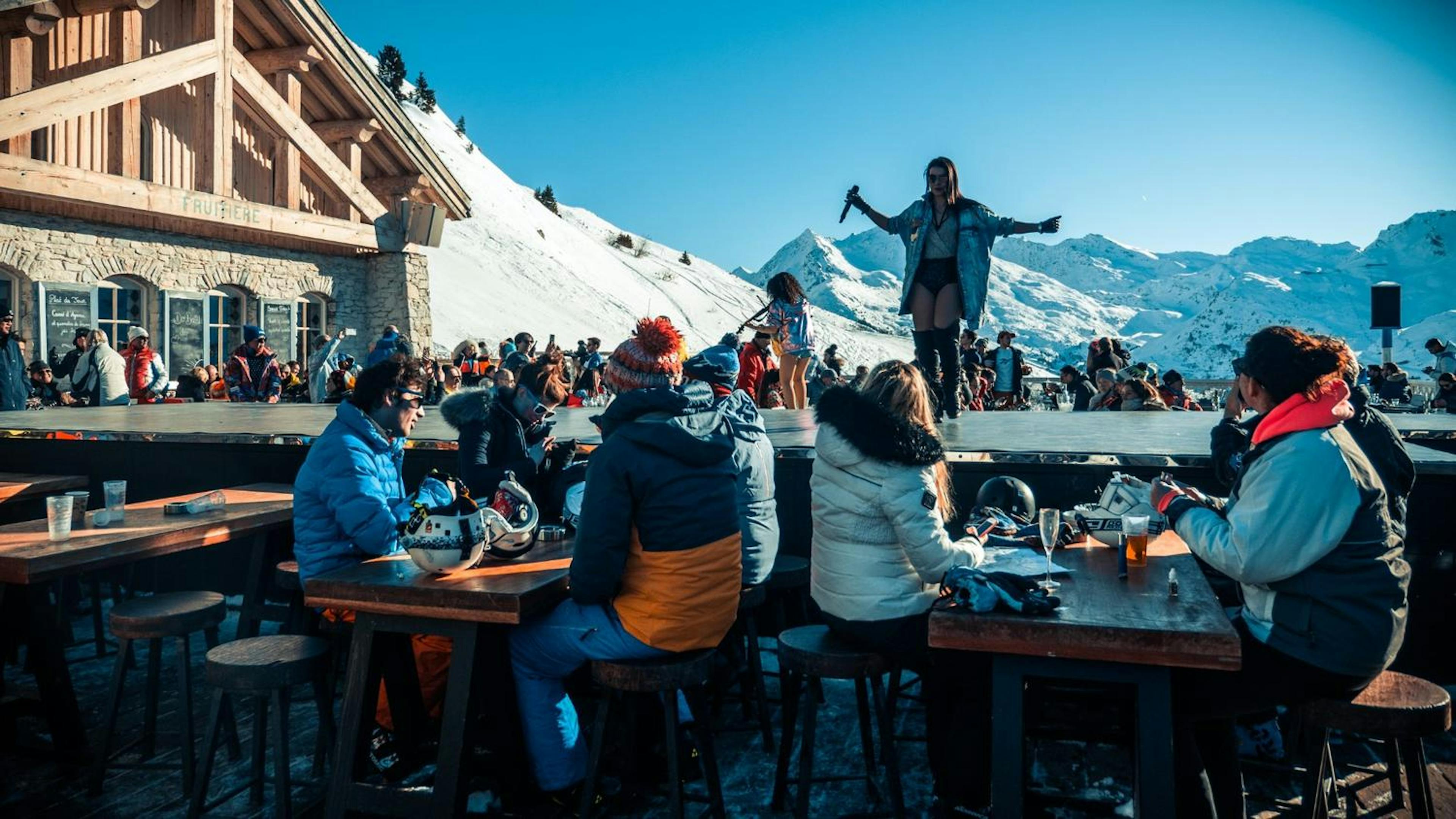 Skiiers watching a performance at an on-mountain patio on a sunny day.