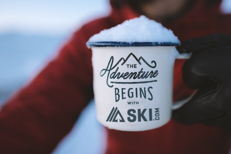 a skier holds a cup of hot cocoa with the logo of ski.com on the cup