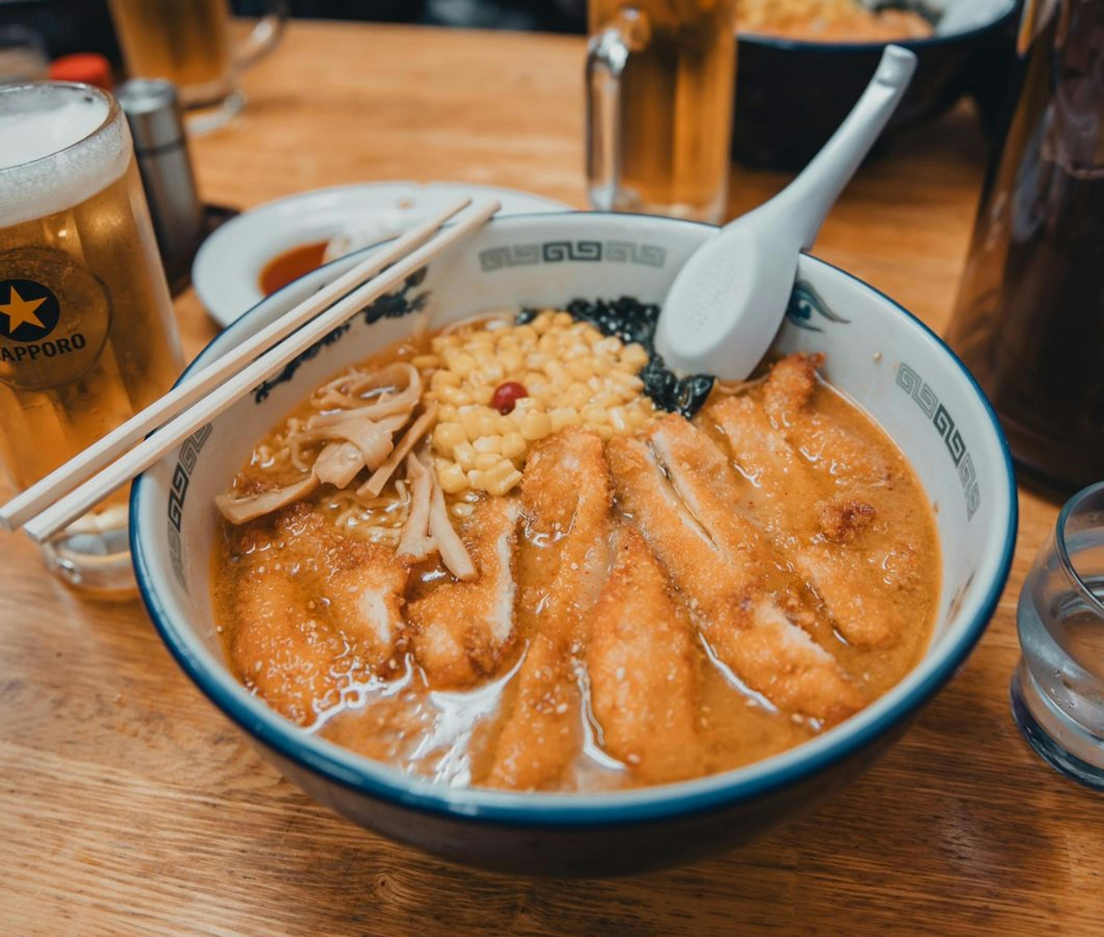 A bowl of steaming ramen accompanied by chopsticks and a cold beer, creating a delightful dining experience.