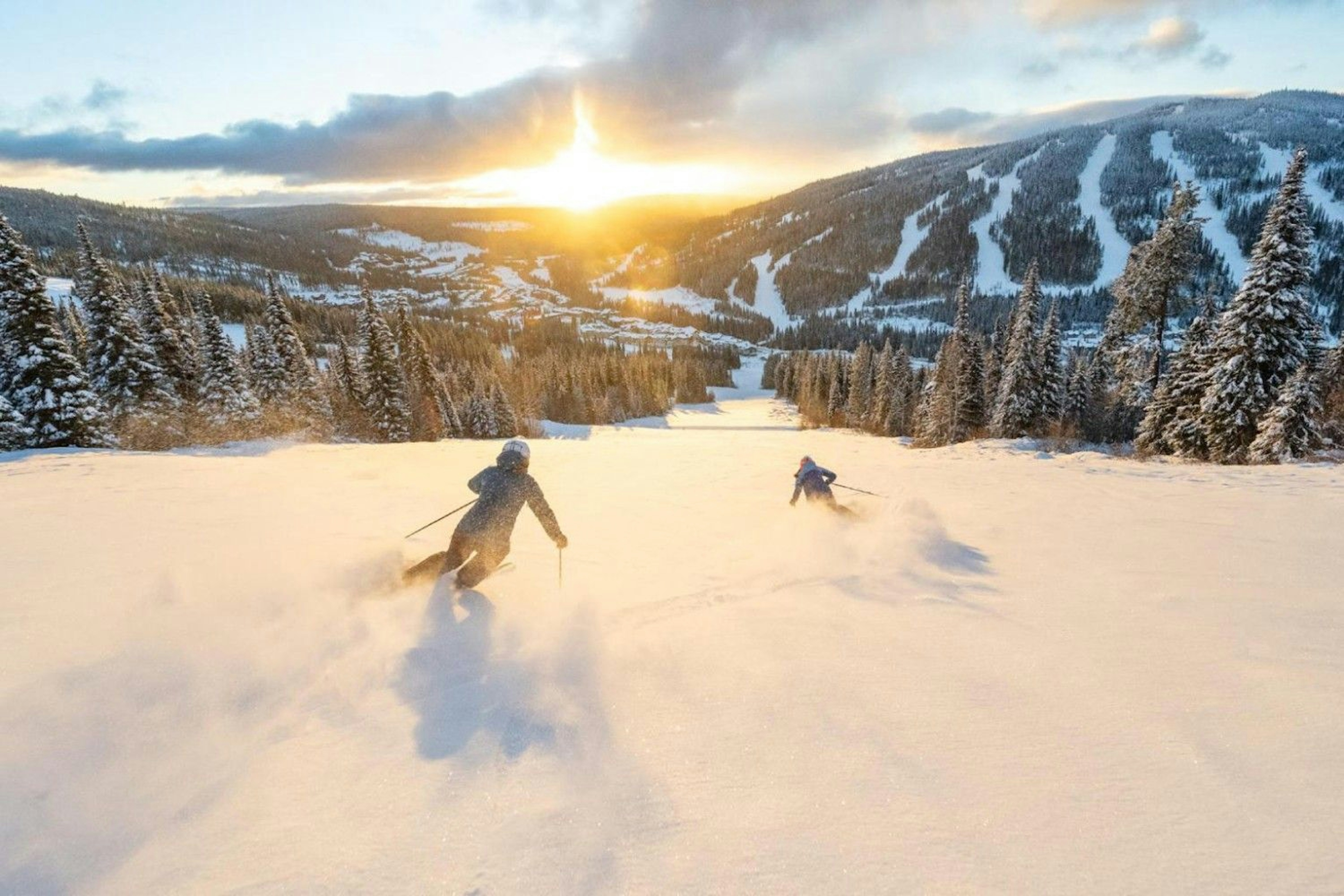 two skiers ride sun peaks resort on a large groomer in the sunset