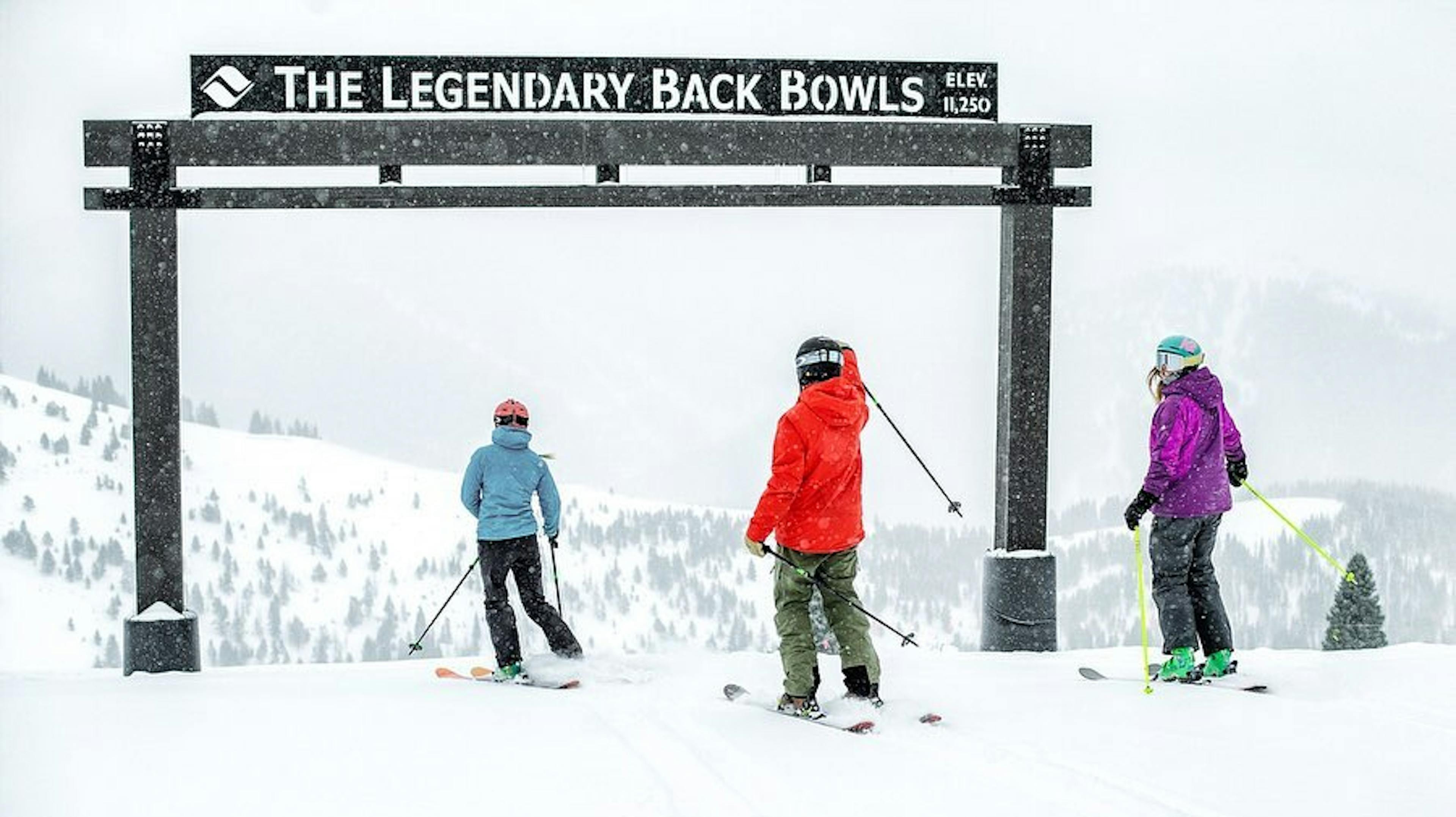 3 skiers stand under The Legendary Back Bowls sign on a snowy day at Vail Mountain Resort