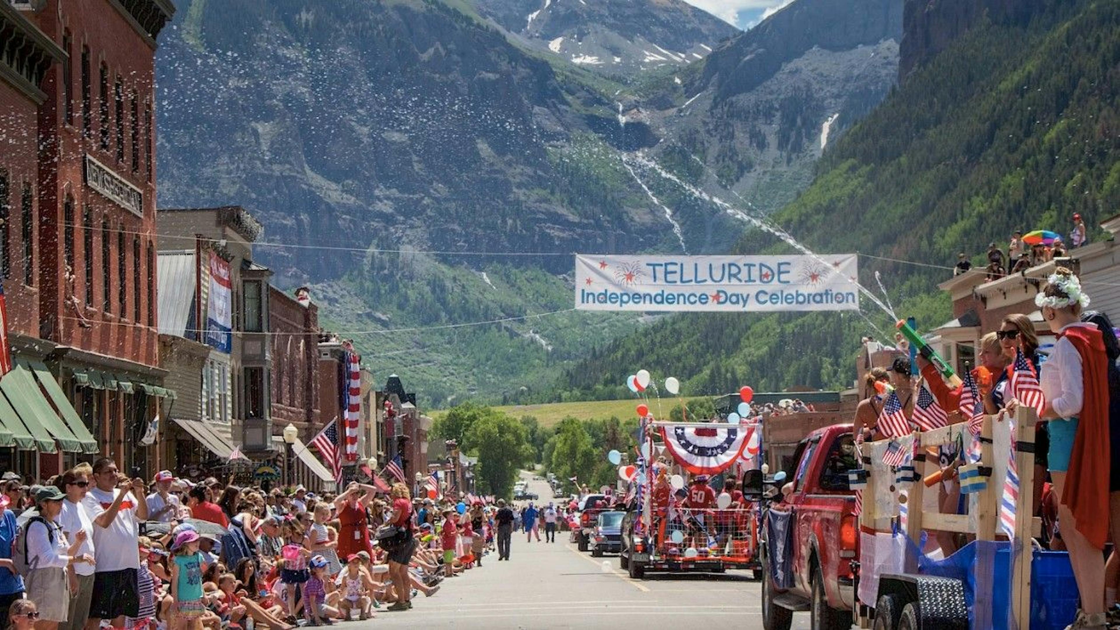 A crowd of people dressed in red, white and blue watch the Telluride 4th of July parade with alpine mountains in the backdrop