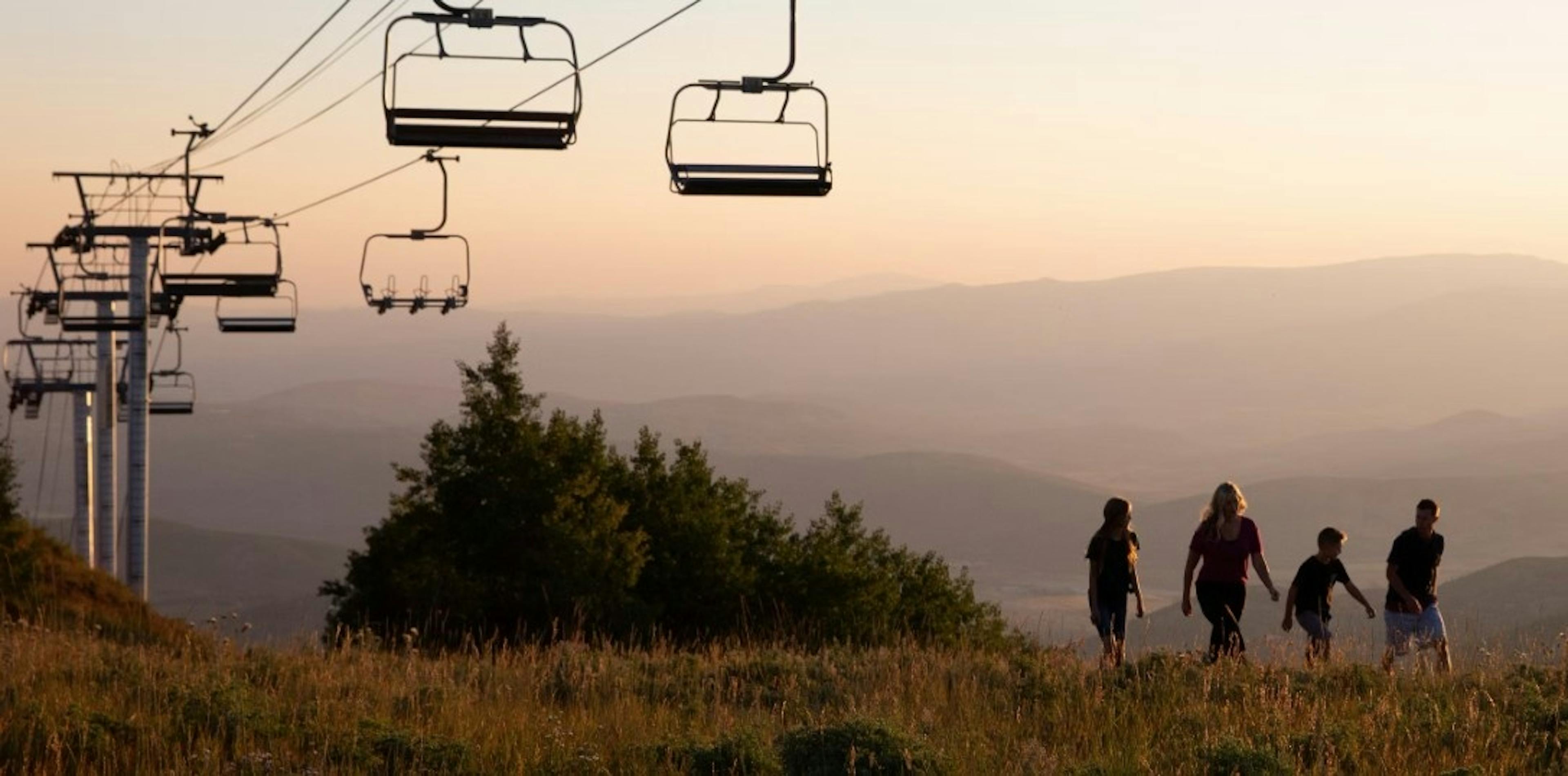 Parents and their 2 kids walk in a grassy field underneath the Park City Mountain Resort ski lift with the sunset and mountains in the backdrop