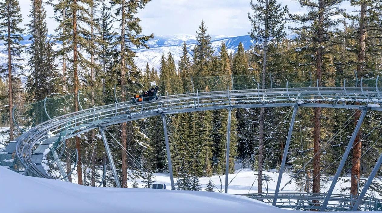 A parent and child ride the Snowmass Breathtaker Alpine Coaster through pine trees and a snowy mountain backdrop