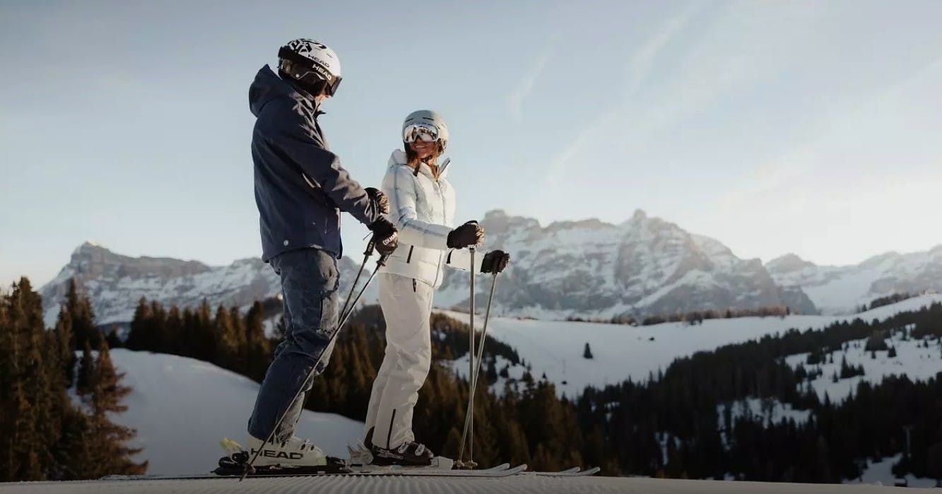 Two skiers are smiling as they stand still on a groomed run at Alta Badia Resort with snowy mountains in the backdrop
