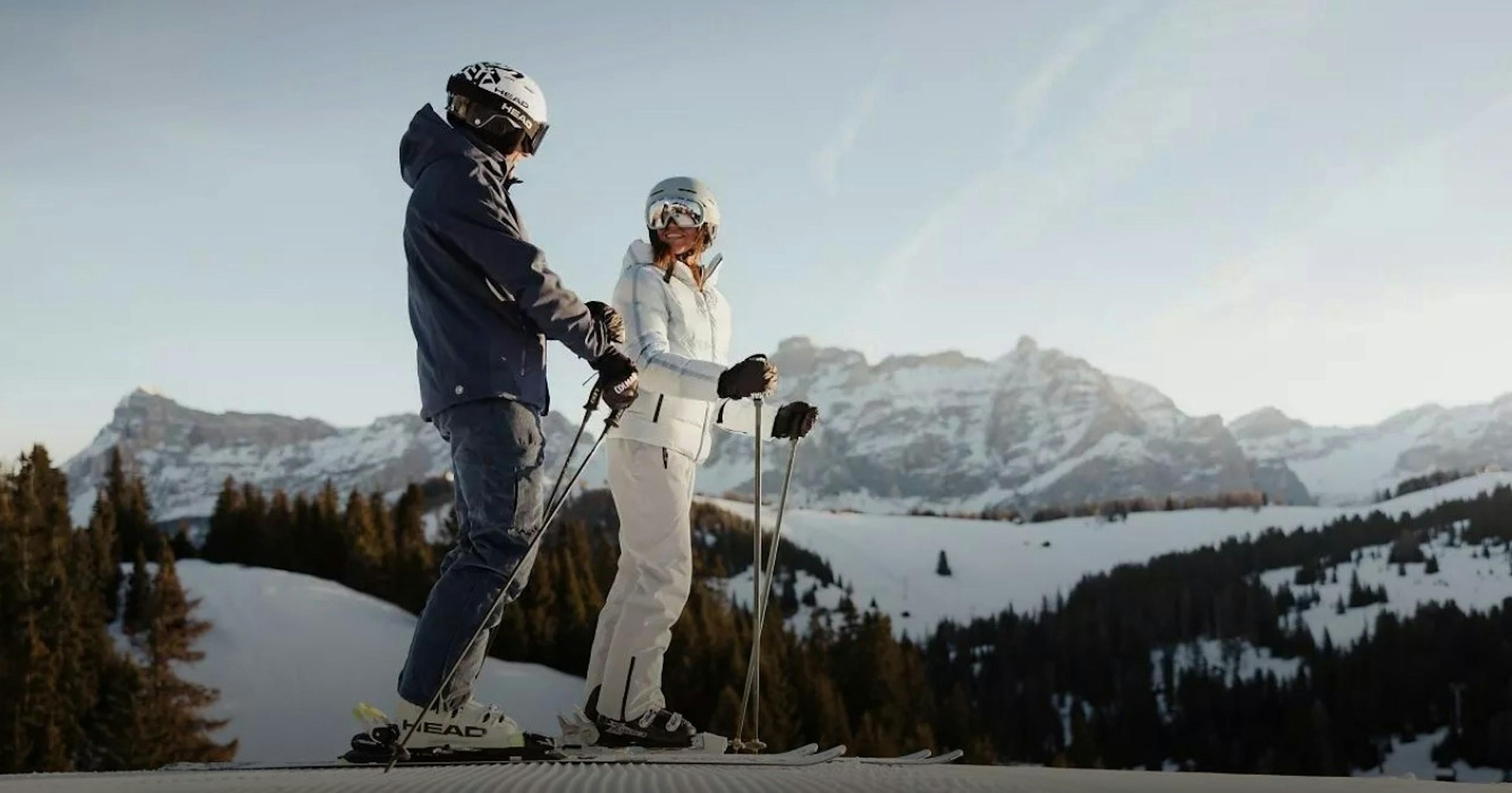 Two skiers are smiling as they stand still on a groomed run at Alta Badia Resort with snowy mountains in the backdrop