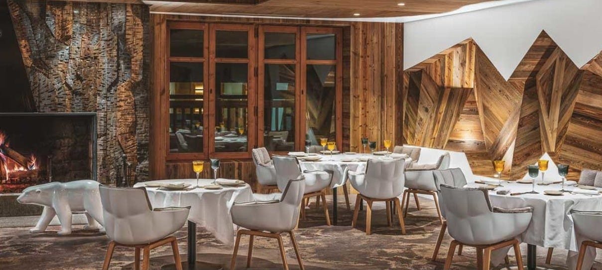 Modern white sofa chairs surround tables with white tablecloths next to a roaring fireplace in the wooden-walled dining room of La Table de l’Ours in France