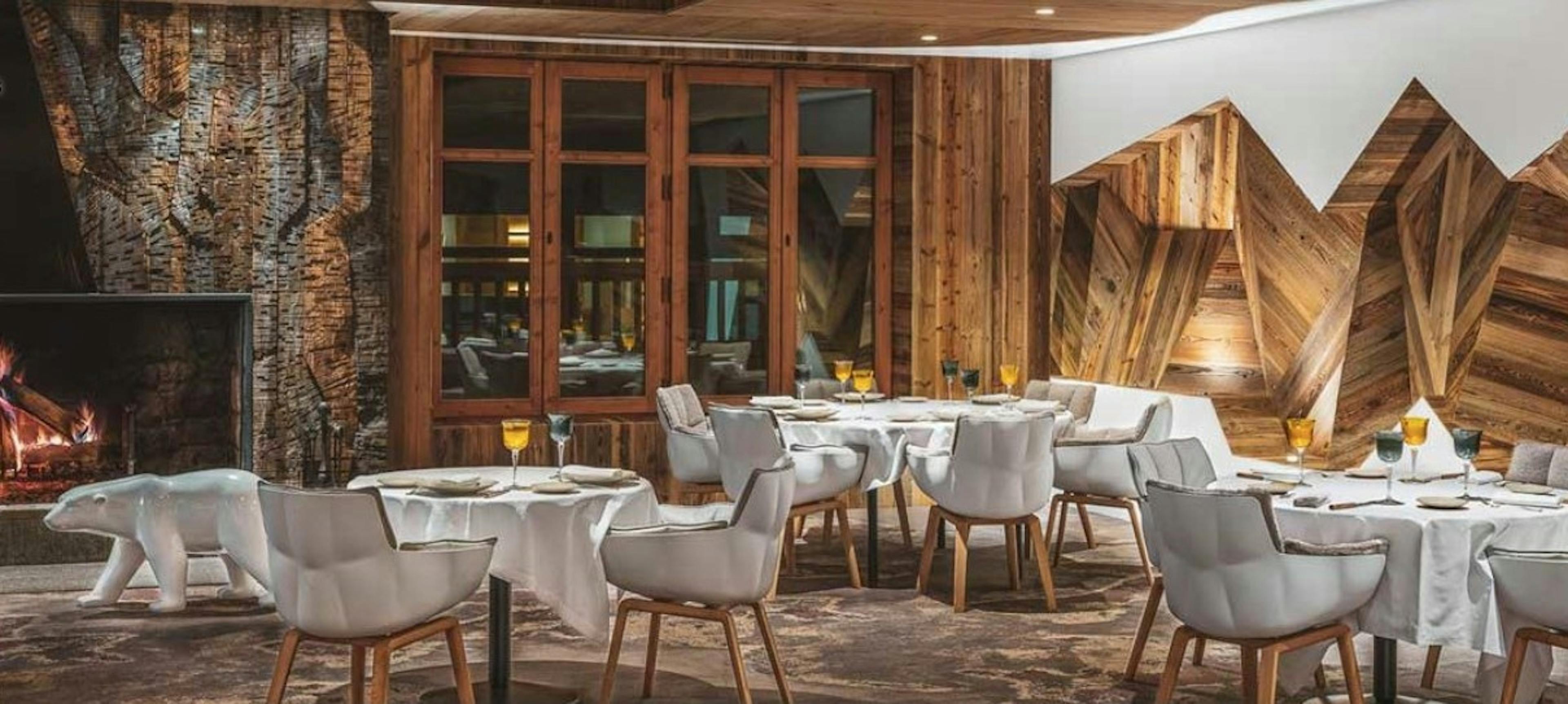 Modern white sofa chairs surround tables with white tablecloths next to a roaring fireplace in the wooden-walled dining room of La Table de l’Ours in France