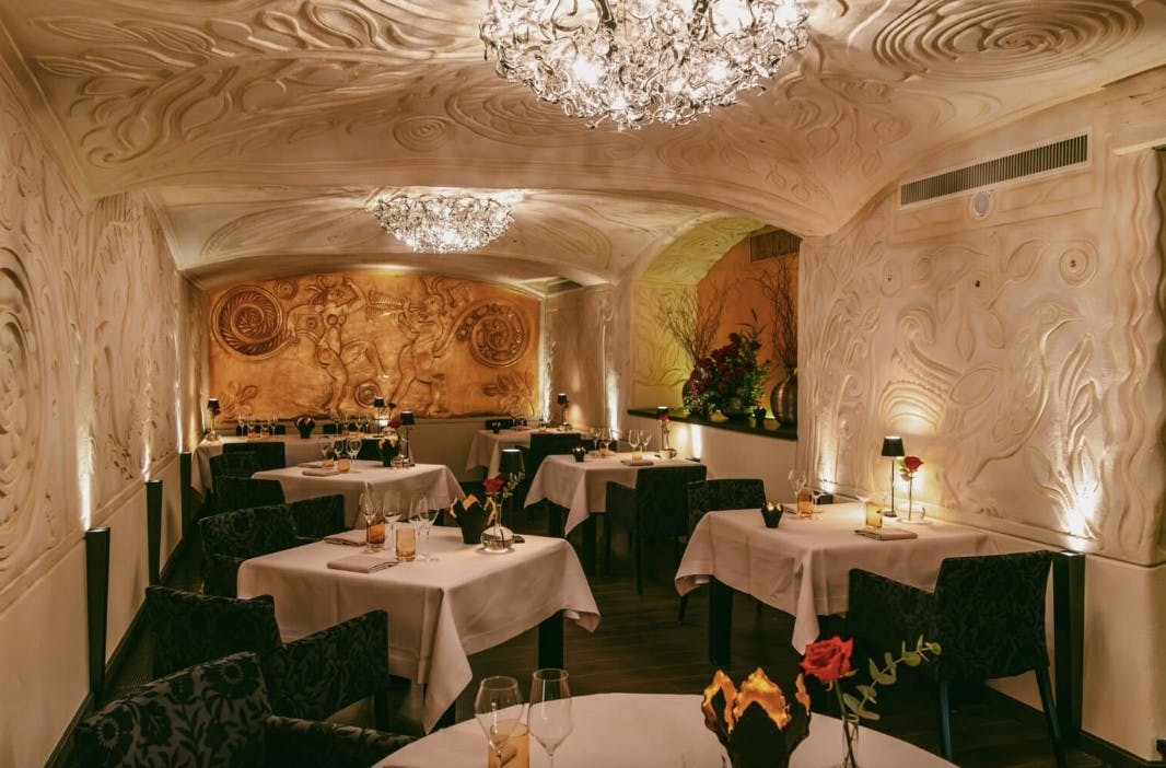Black wooden chairs surround 7 tables with white tablecloths and crystal chandeliers in the fine dining room of Ecco St. Moritz