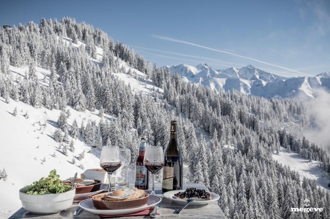 A table with French cuisine and 2 glasses of red wine with a backdrop of snowy alpine trees and mountains