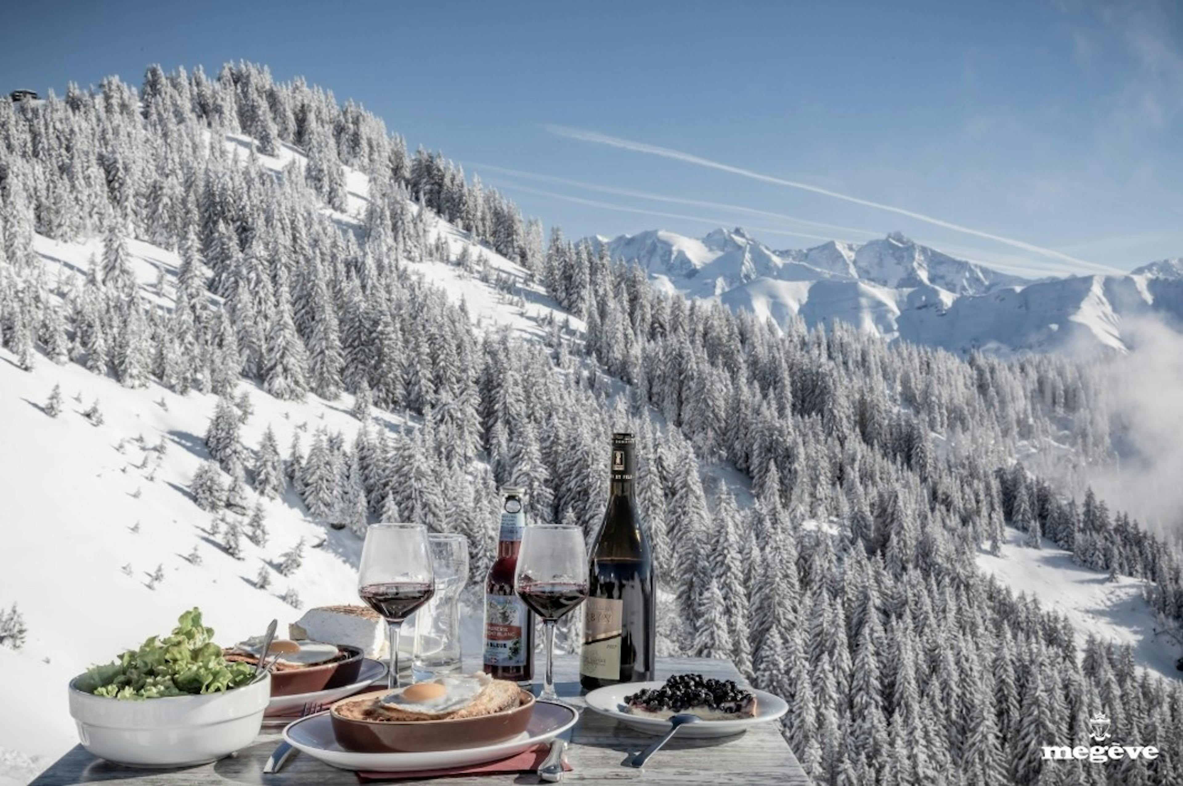 A table with French cuisine and 2 glasses of red wine with a backdrop of snowy alpine trees and mountains