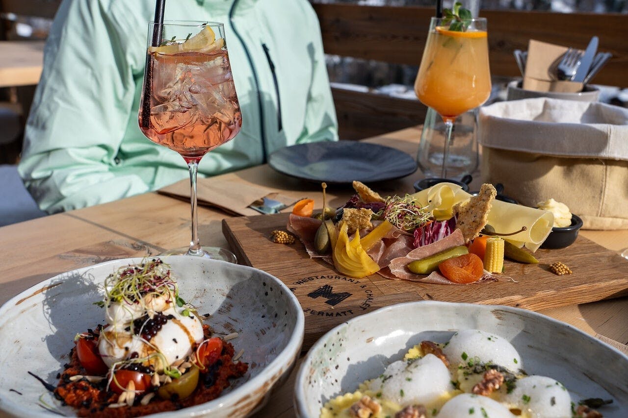 Plates of fine dining cuisine and wine glasses sit on a sunny patio table in Zermatt