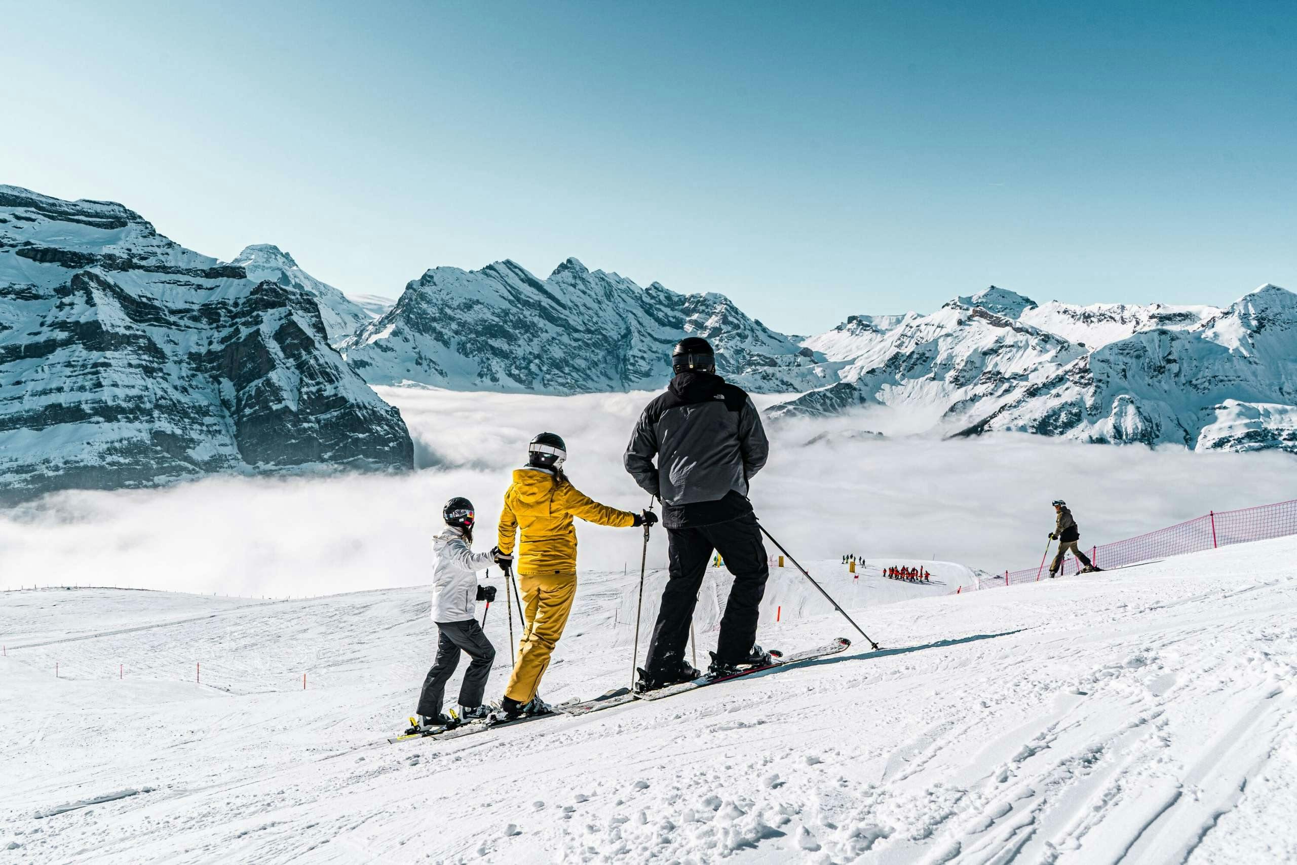 Male ski guide in black and grey ski attire tending to two young student skiers on the slopes of Wengen, Switzerland.
