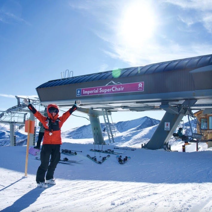 Person in ski patrol outfit pumps their arms in the air standing in front of the Imperial Express SuperChair at Breckenridge Resort
