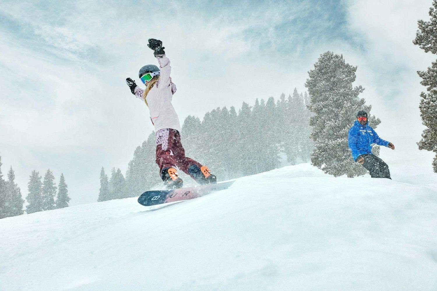 Child snowboarder cheers with arms up as they conquer a jump with a Breckenridge ski school instructor