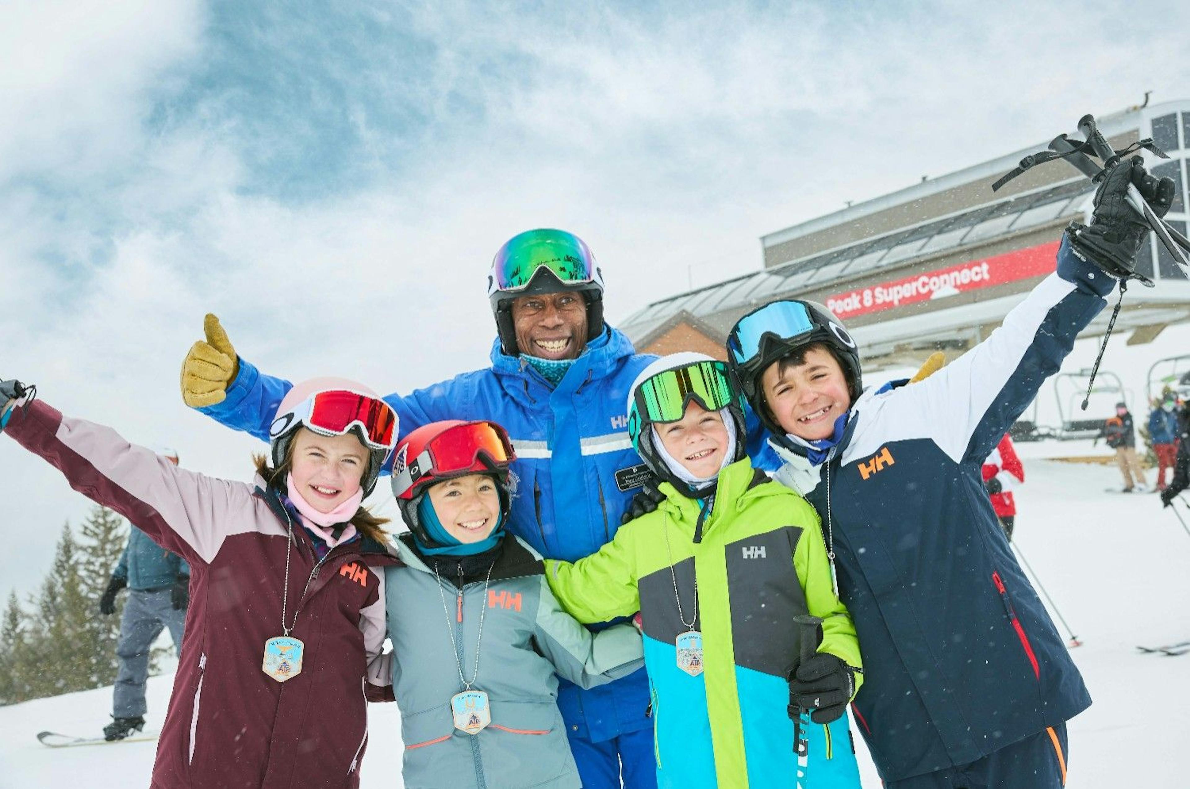 A Breckenridge ski school instructor and four kids in ski gear smile and pose for a photo in front of a ski lift at Breckenridge Ski Resort
