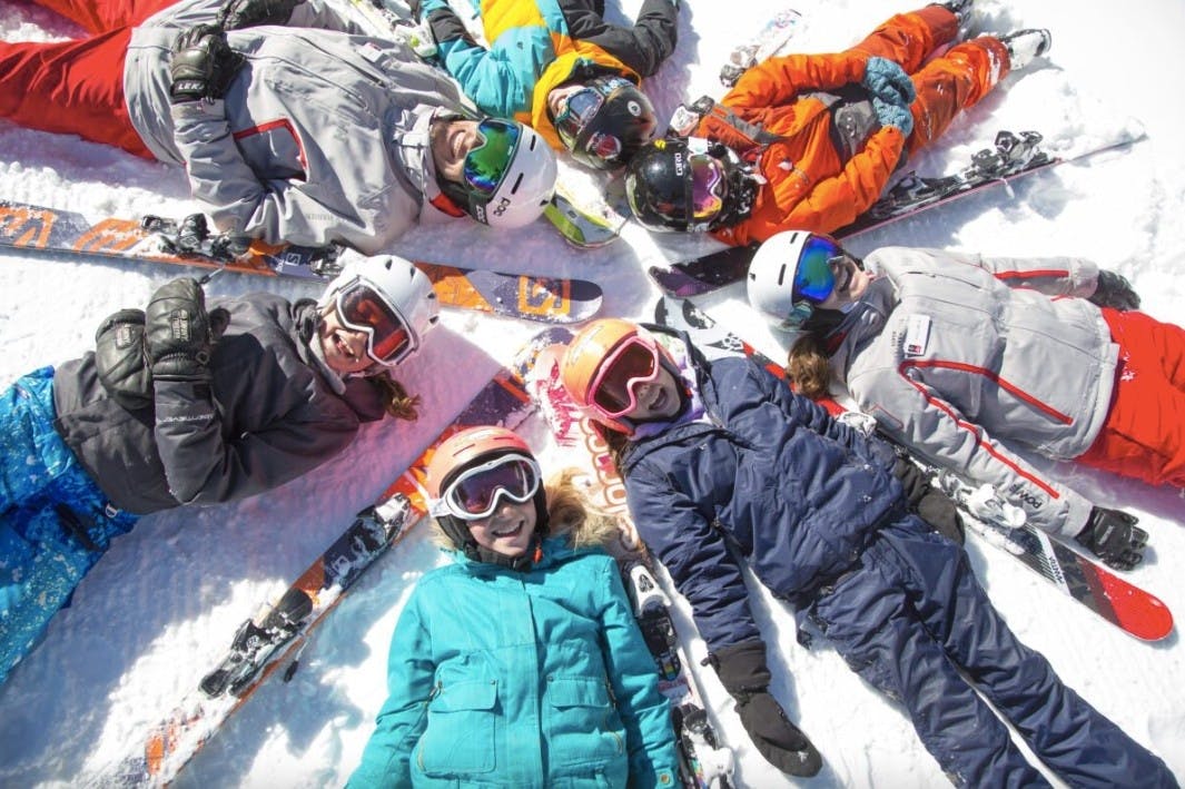 A group of teens in ski gear lie smiling on the ground in the snow in a circle