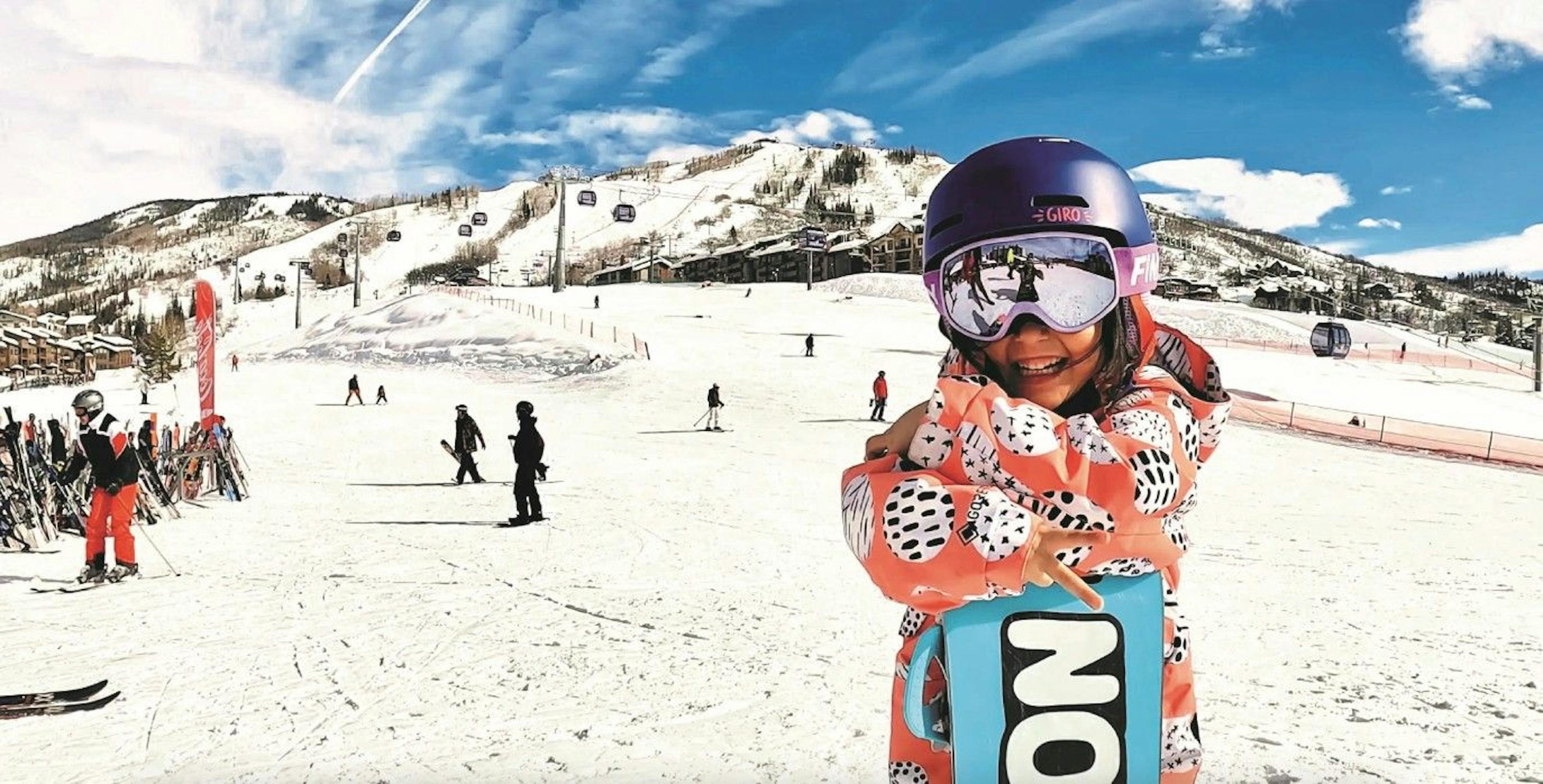 A small child in snowboarding gear smiles and gives the peace sign with their fingers, standing at the bottom of the Steamboat Springs Resort mountain on a bluebird day