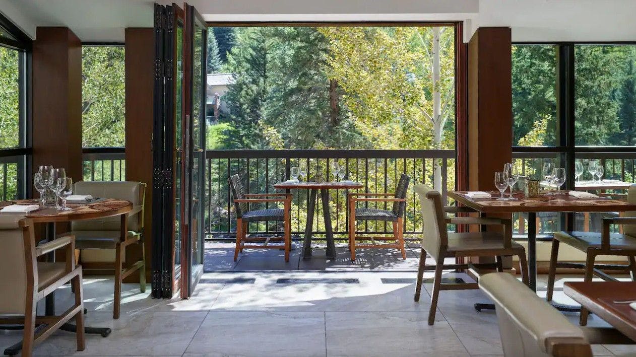 The modern dining room of Gessner Vail restaurant with set tables and chairs and open doors to a patio with creekside views