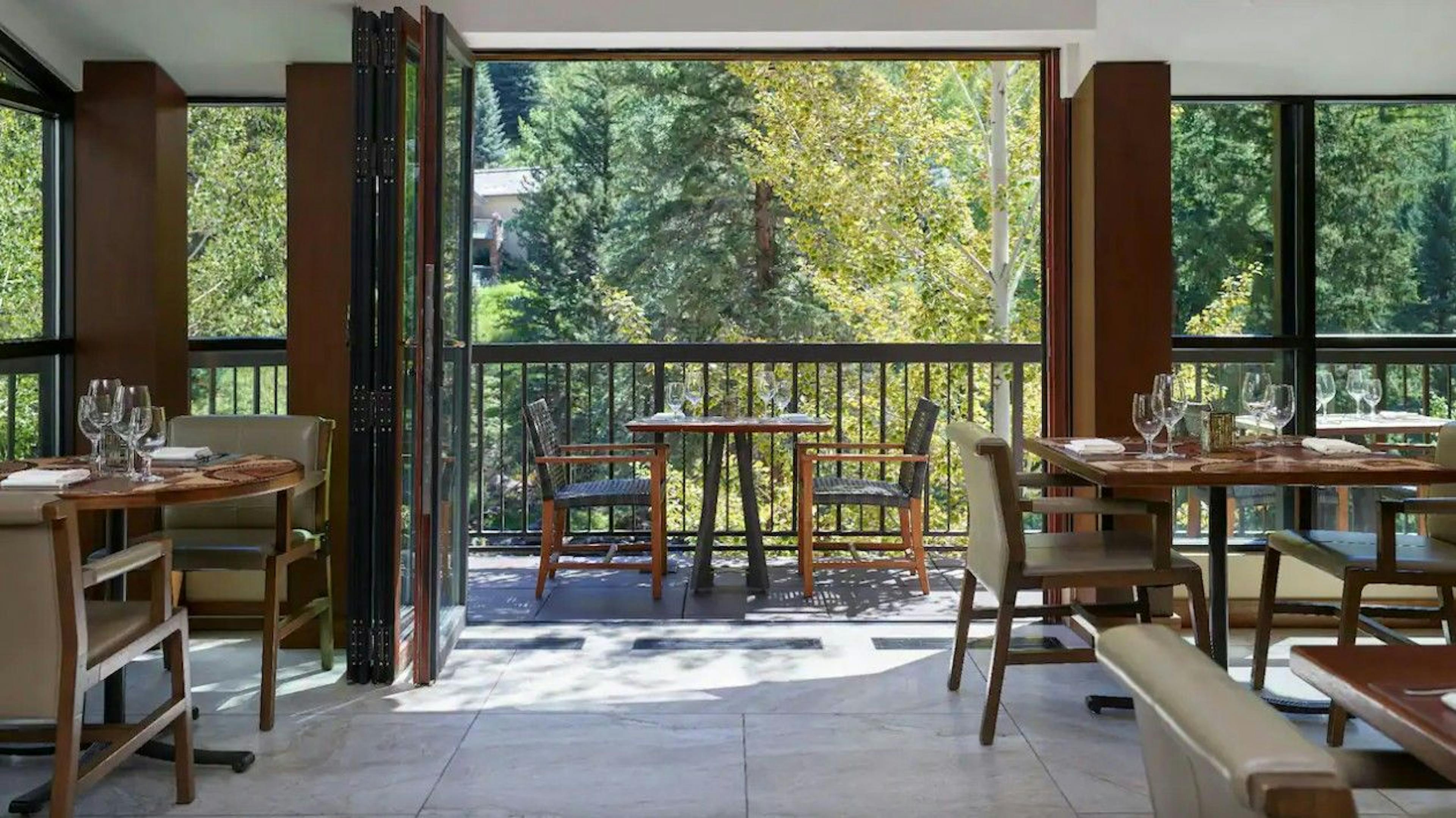 The modern dining room of Gessner Vail restaurant with set tables and chairs and open doors to a patio with creekside views