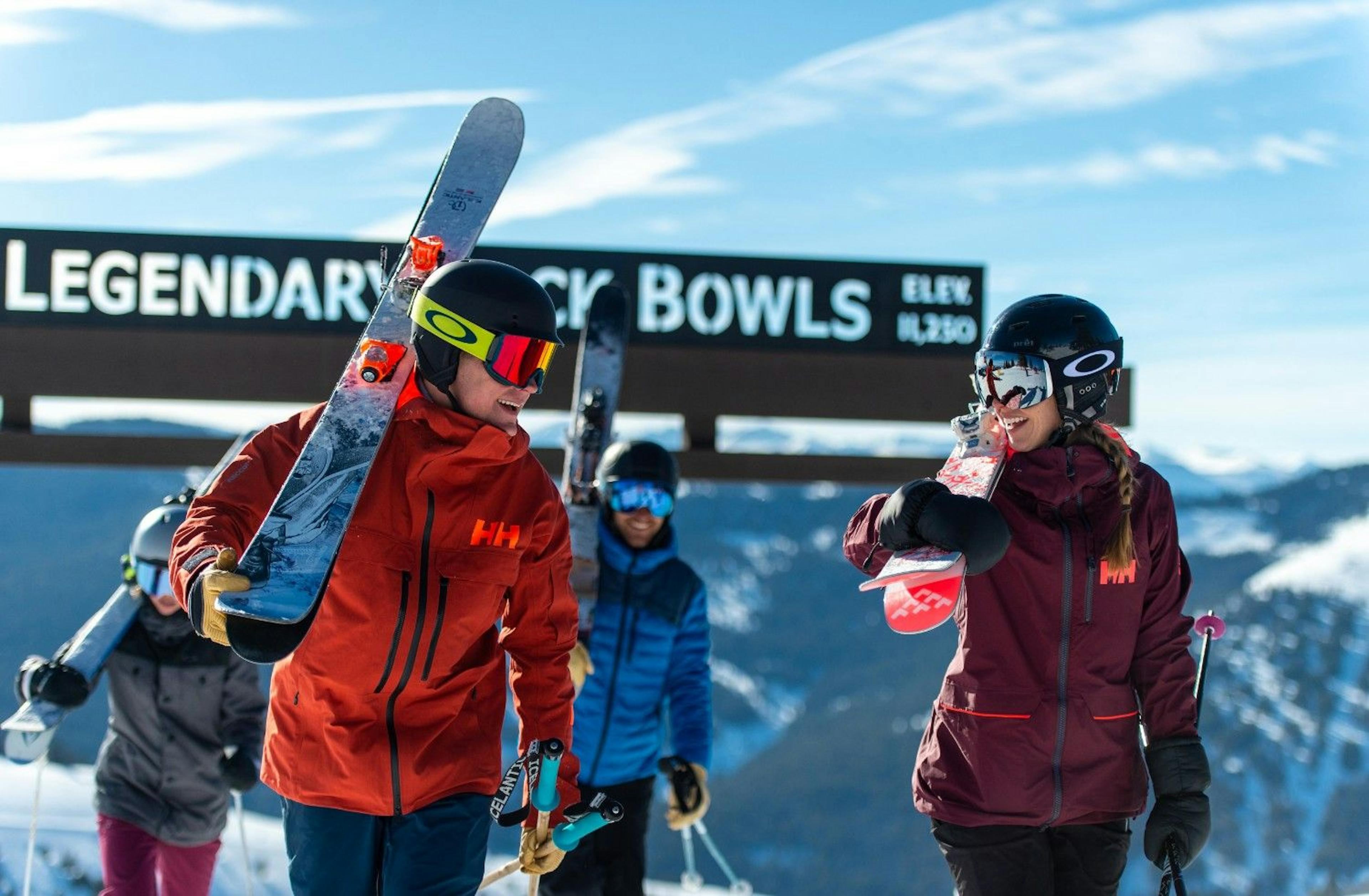 four skiers carrying their skis and smiling as they walk away from the Legendary Back Bowls sign at Vail Resort