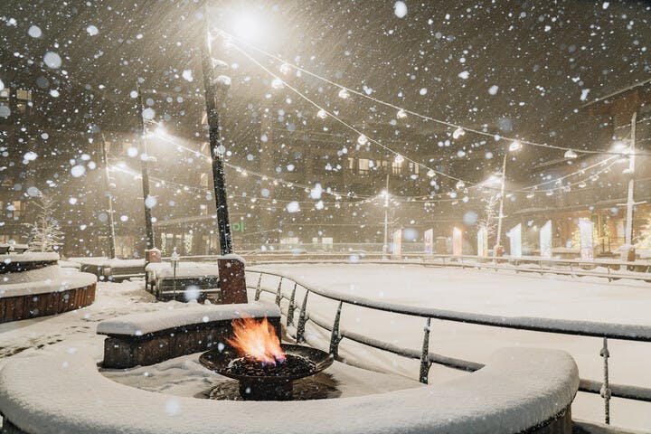 snow falls on a patio with a fire pit framed by twinkling string lights