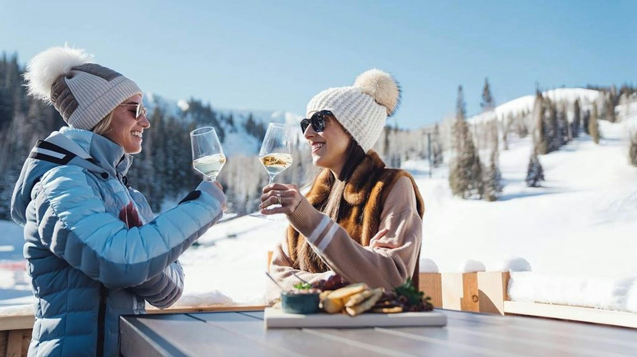 two smiling women wearing winter clothes cheers their white wine glasses on a sunny porch with the Deer Valley ski slopes behind them 