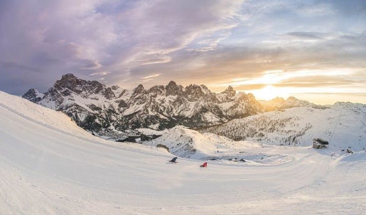 The sun sets over the snowy mountains and slopes of Cortina d'Ampezzo as two people ski down the run