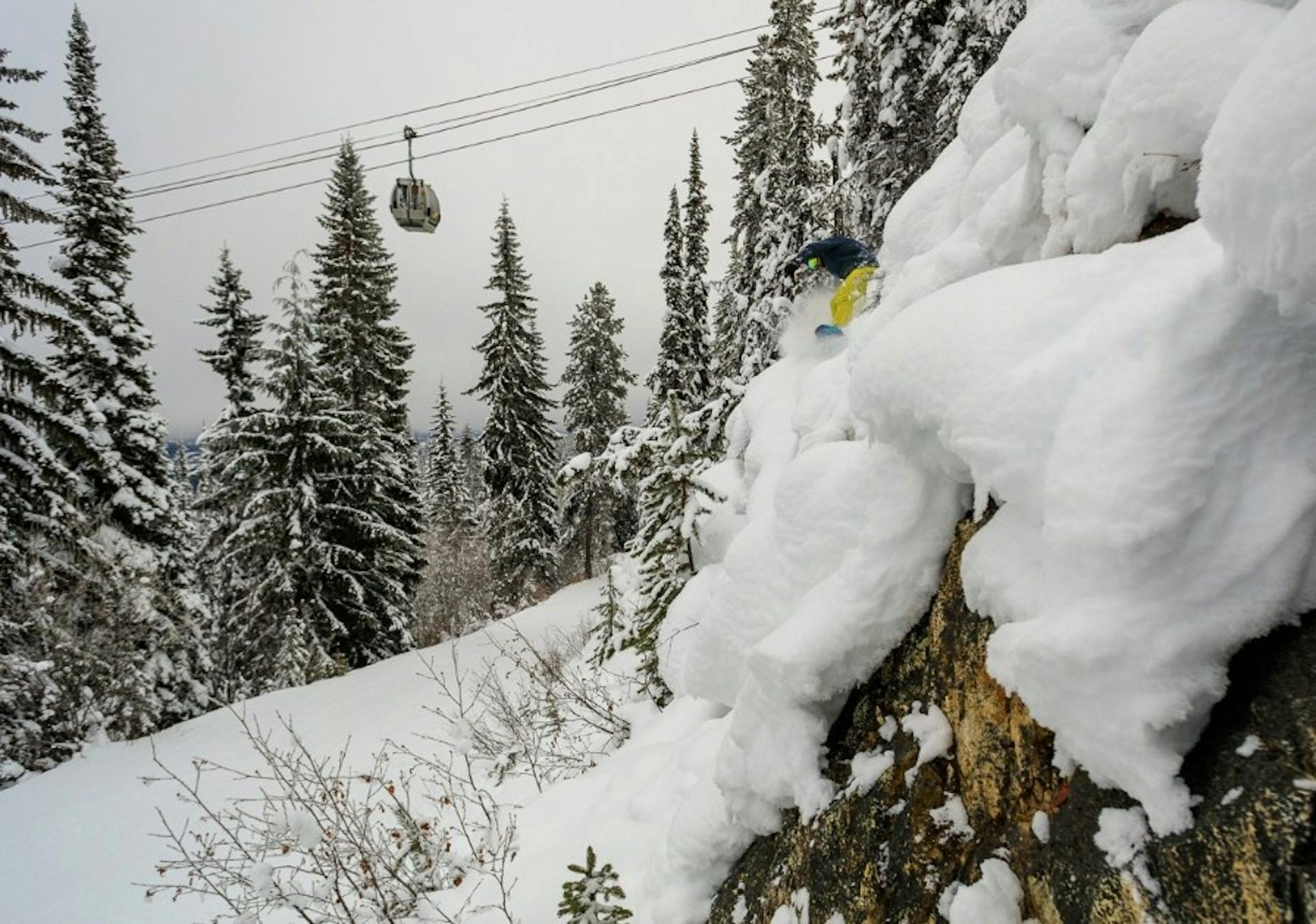 Snowboarder sending it down a steep cliff in some expert terrain at Kicking Horse Resort with the lift spinning behind them