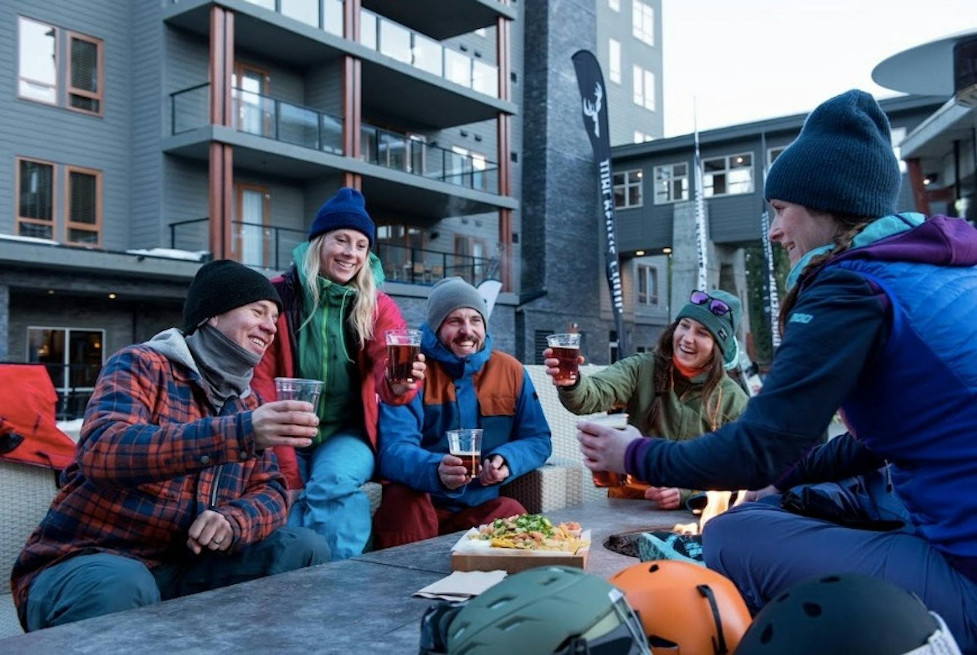 Group of friends sitting on a patio laughing and clinking cups of beer as they share a pizza around a fire pit