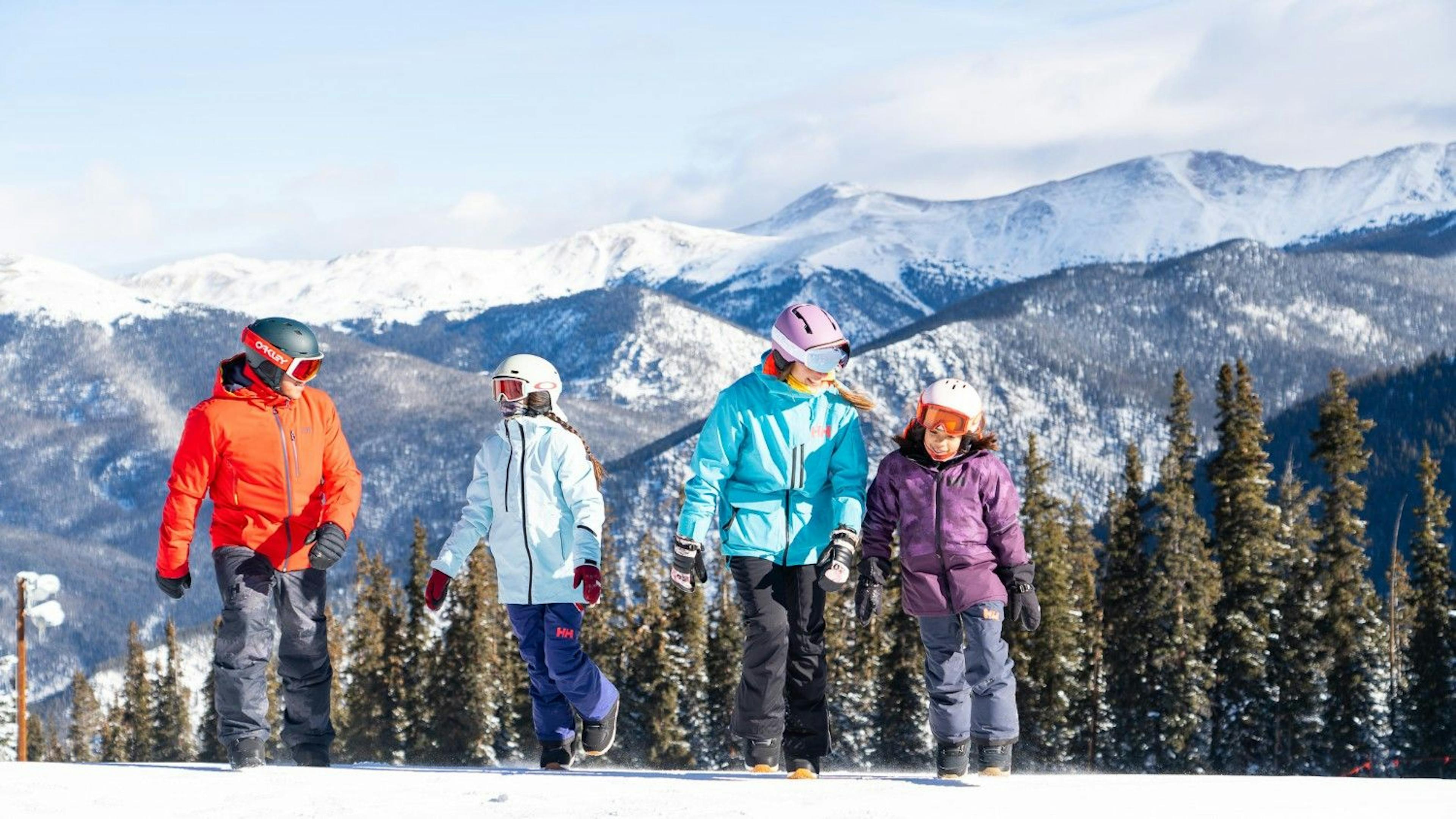 Family of four in snow gear talking and walking on the snow with snowy mountains behind them