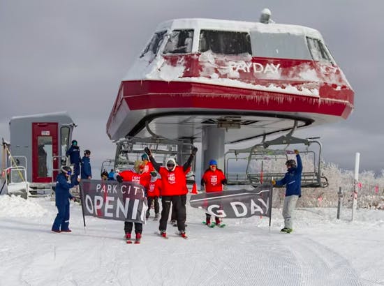 With arms in the air, skiers ride through and break a sign that says Park City Opening Day at the top of a ski lift at Park City Resort