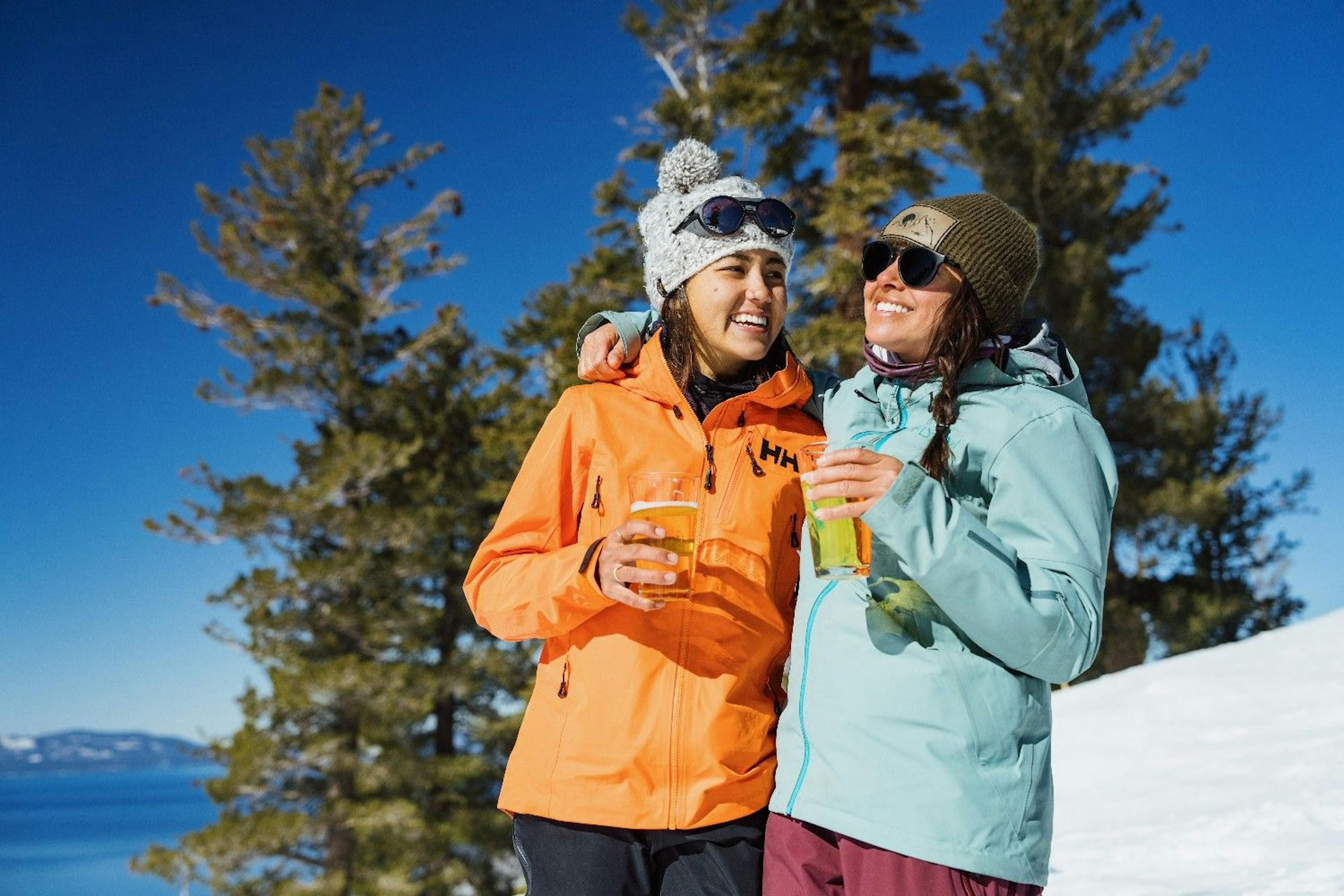 Two friends in snow gear smile and laugh as they drink beers on a sunny day at Heavenly Resort overlooking Lake Tahoe
