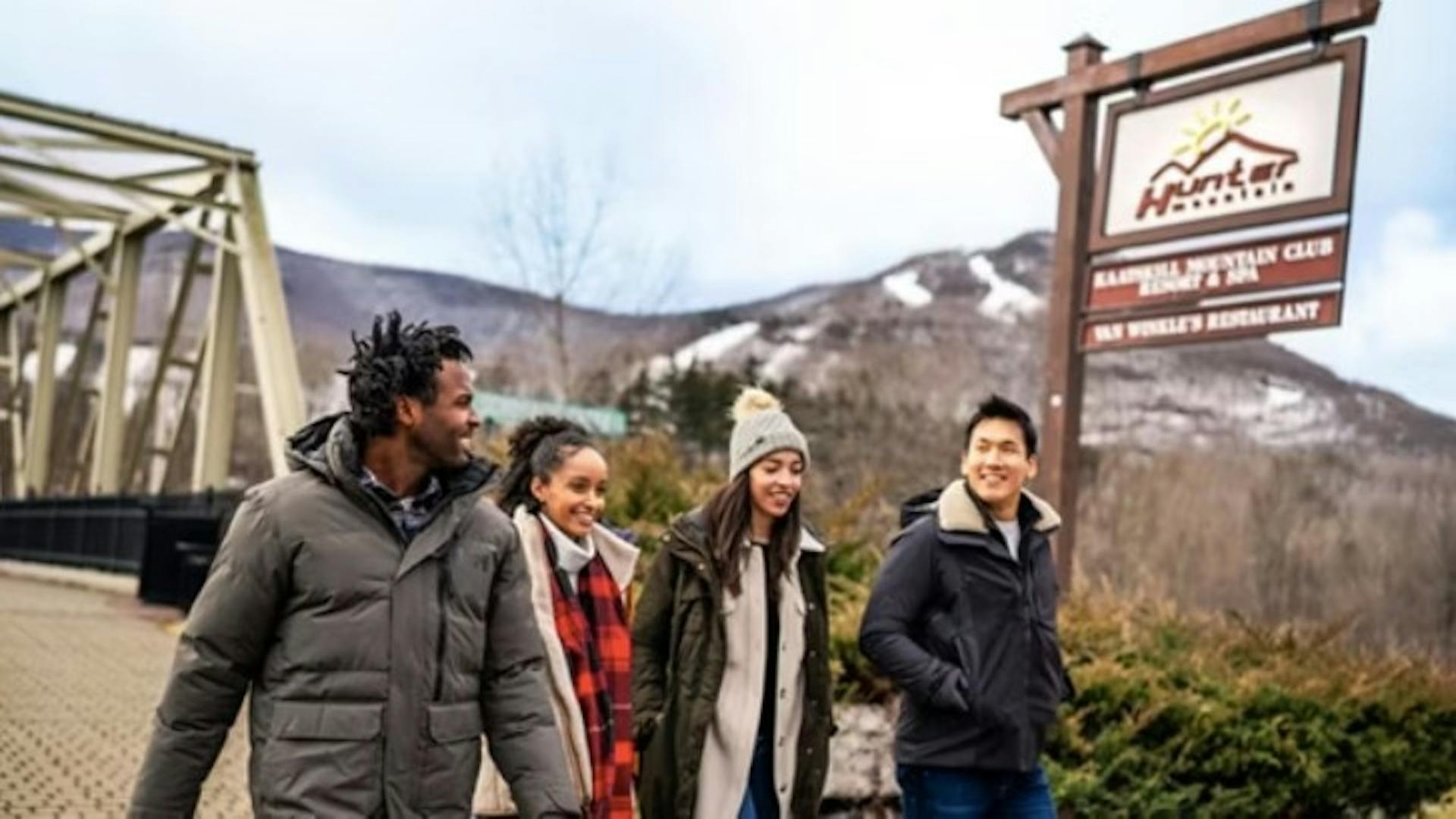 Group of 4 friends in winter clothes walking and smiling in front of a sign and the slopes of Hunter Mountain Resort