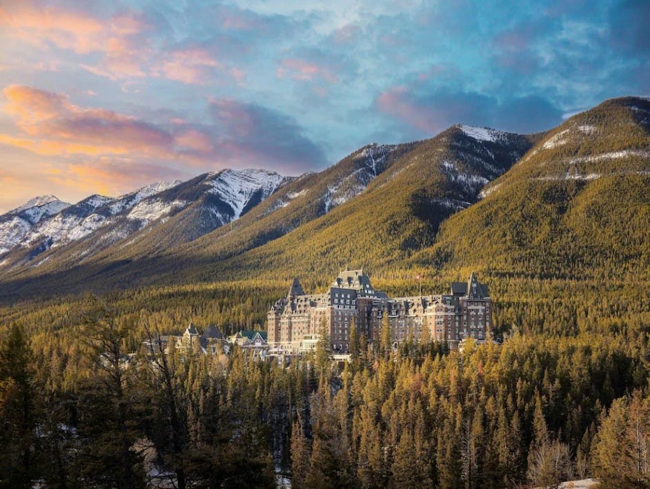 Aerial view of the Fairmont Banff Springs Hotel at sunset with snowy mountains and evergreen trees