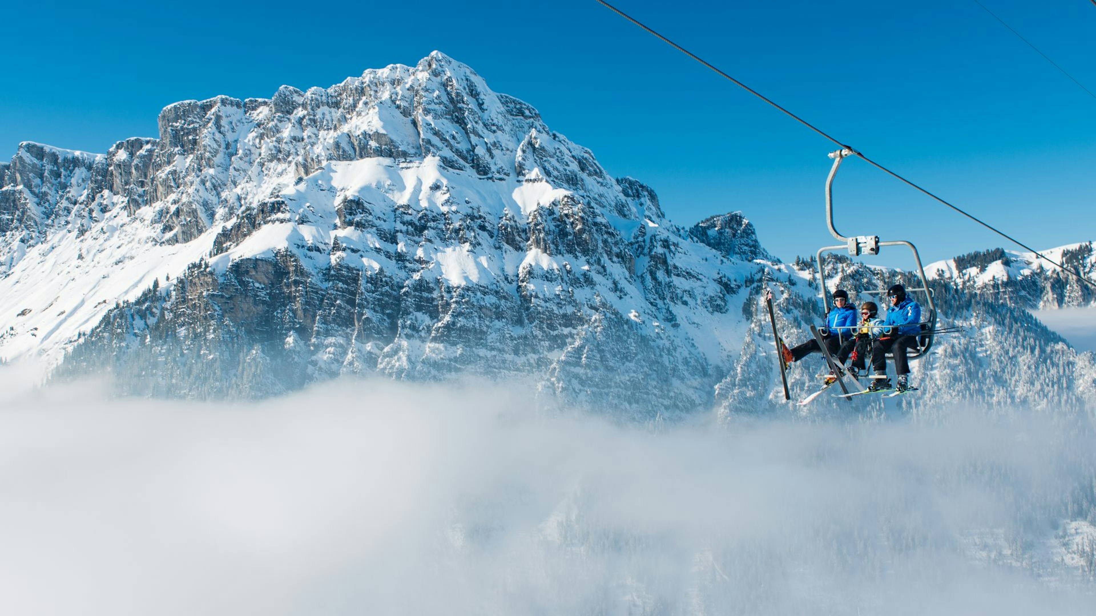 Four people ride a ski lift above the clouds, with a snow-covered mountain peak and blue sky in the background.