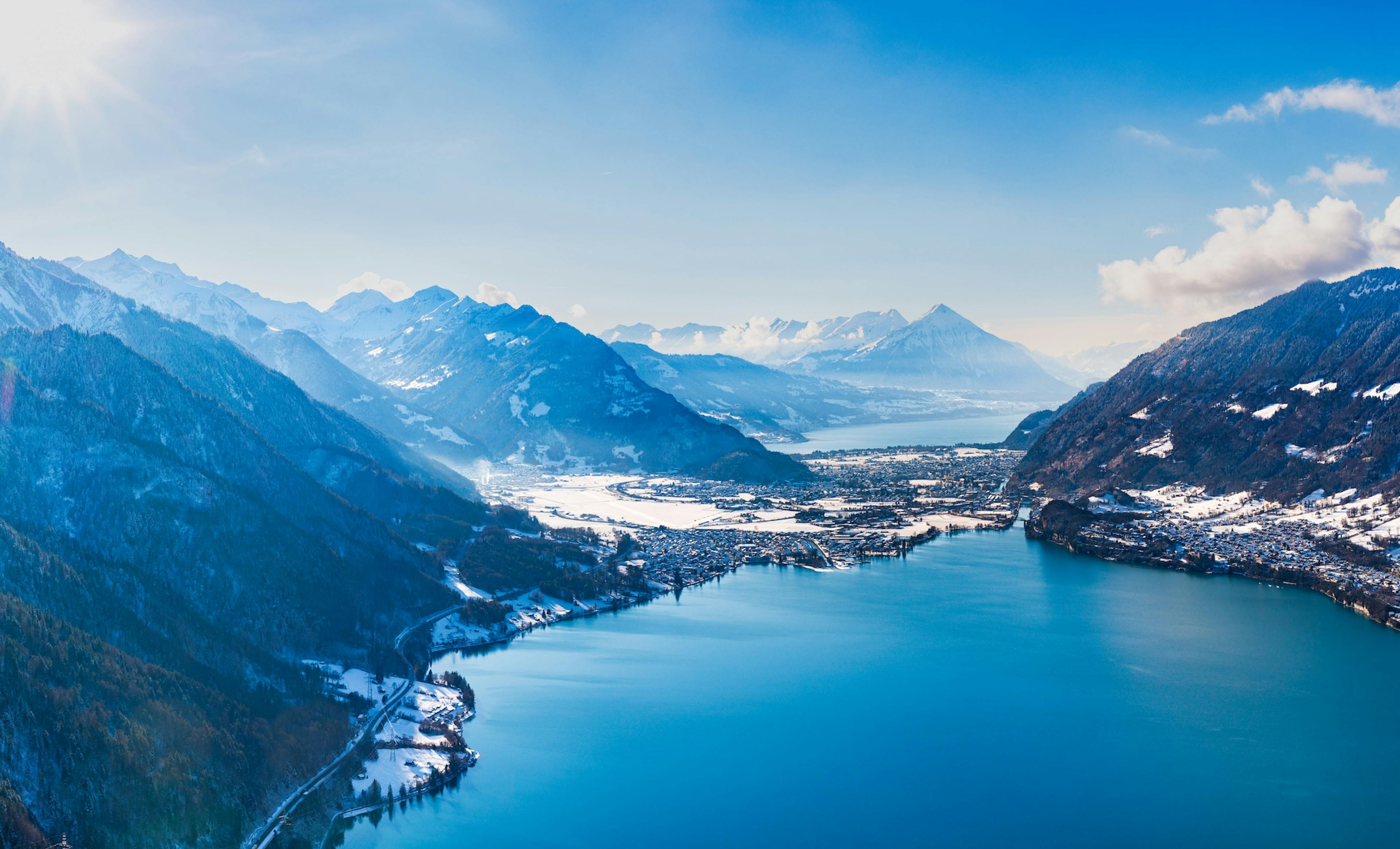 A panoramic view of snow-capped mountains overlooking a tranquil blue lake, framed by a clear sky and sunlight.