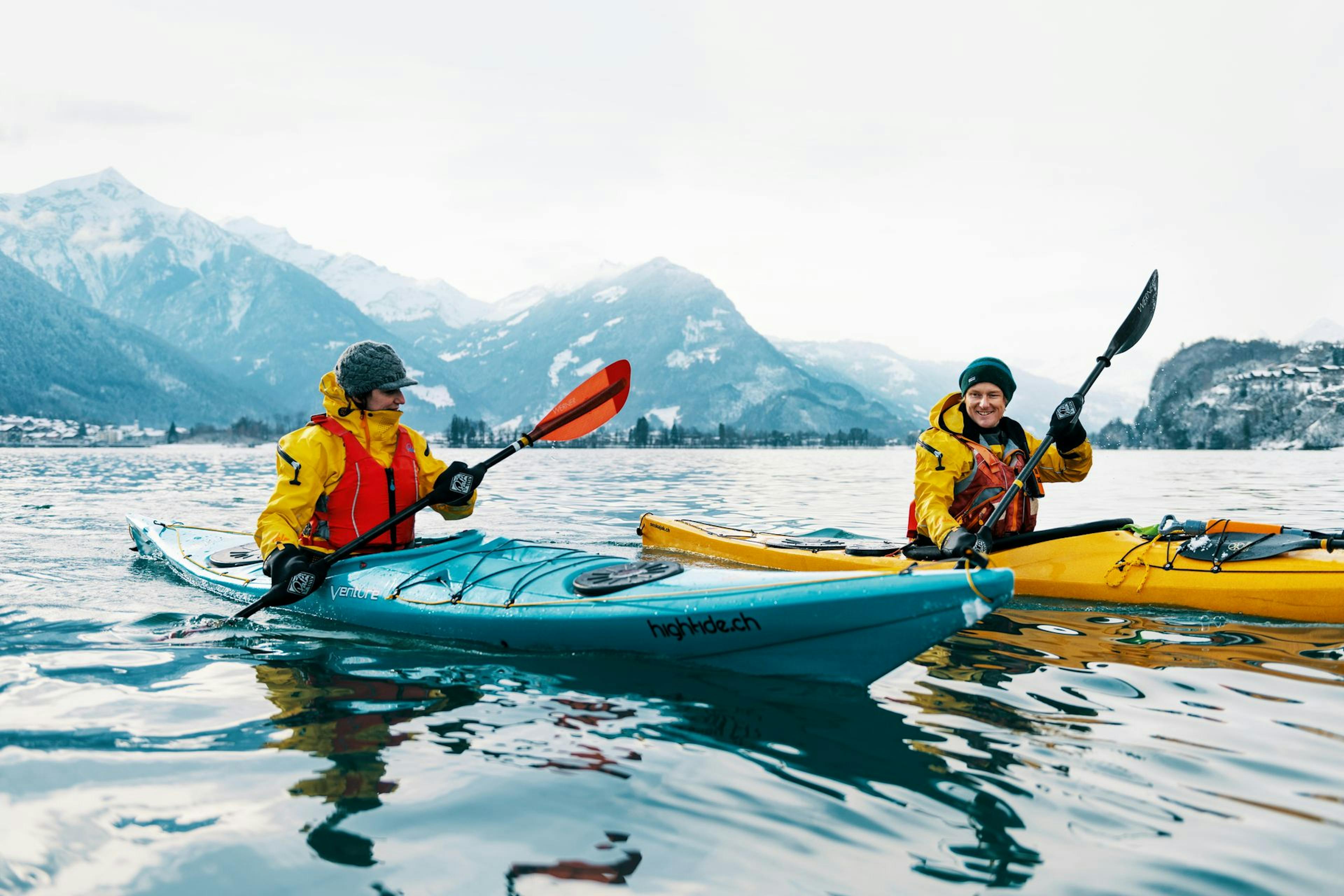 Two kayakers in yellow jackets paddle through calm waters, surrounded by stunning snow-capped mountains under a cloudy sky.