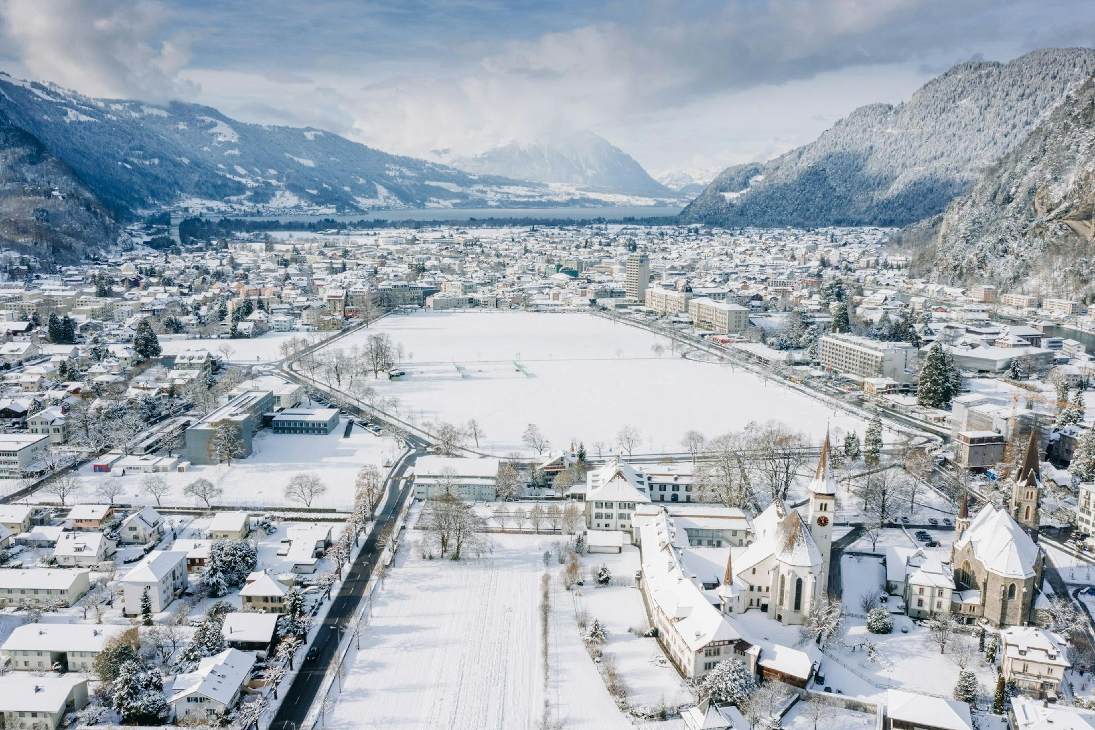 A snowy mountain town with rooftops covered in white, a frozen lake, and distant hills under a cloudy sky.