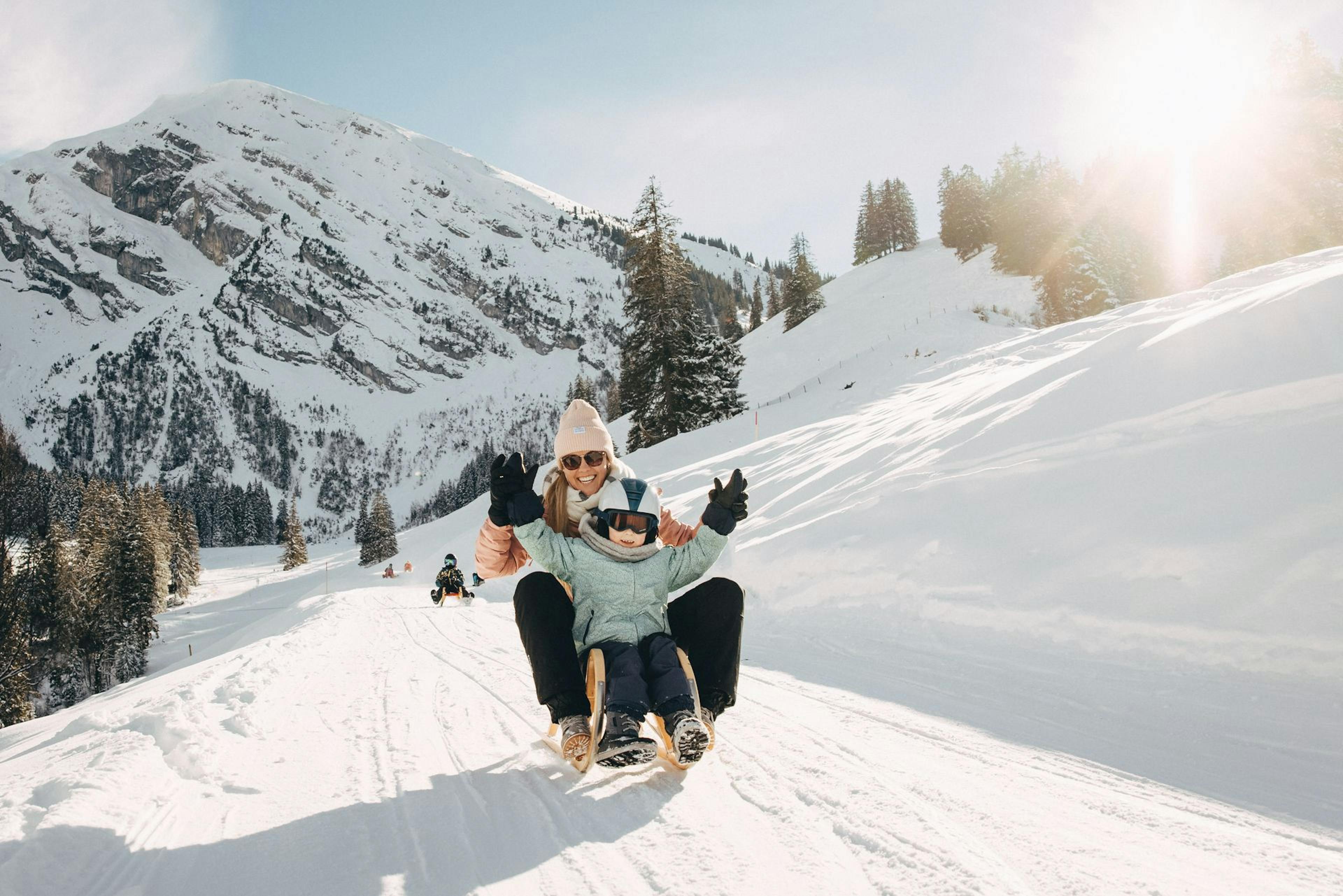  A child wearing a winter coat and helmet joyfully rides a sled down a snowy slope, surrounded by mountains and pine trees.