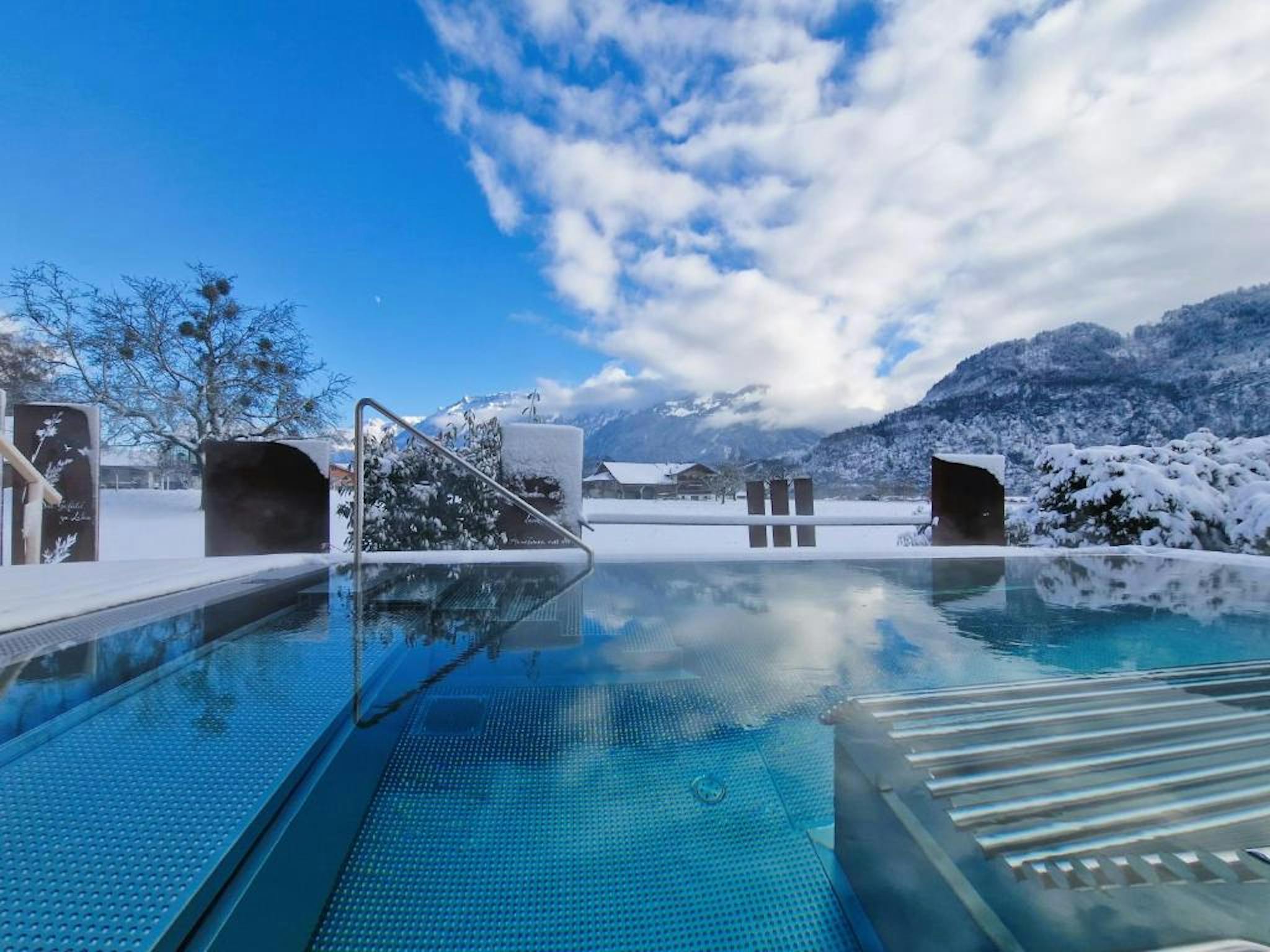 Outdoor infinity pool in snowy landscape, reflecting a blue sky with fluffy clouds. Snow-covered trees and mountains create a serene, wintery scene.