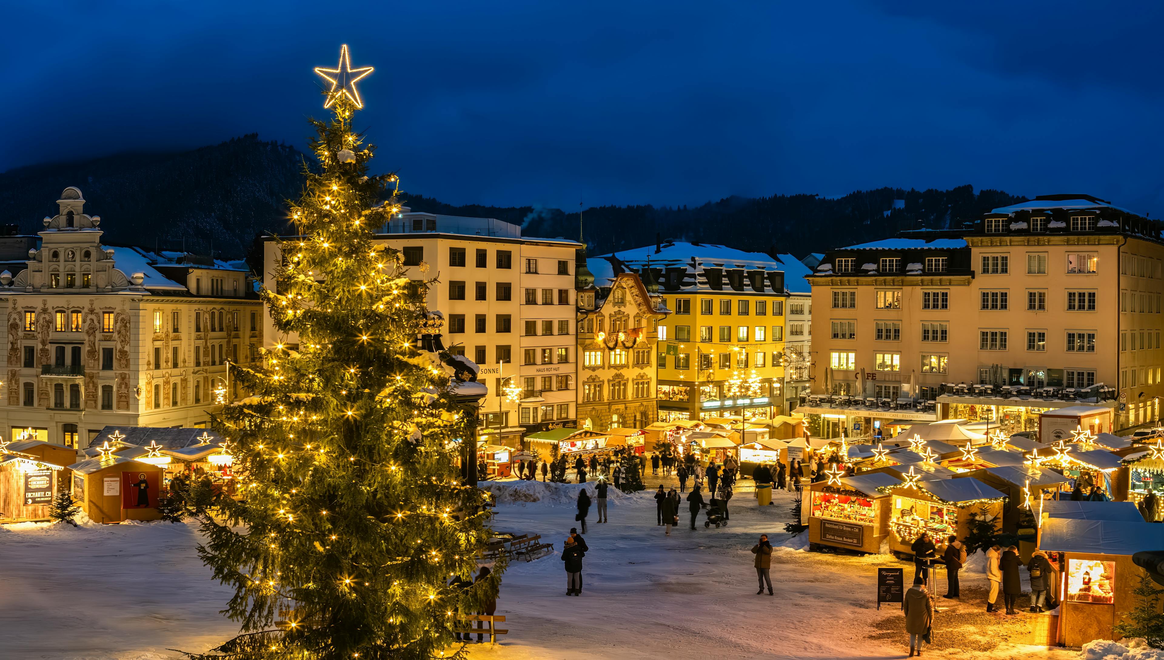 A festive Christmas market scene features stalls, a decorated tree, and crowds enjoying the snowy atmosphere under a twilight sky.